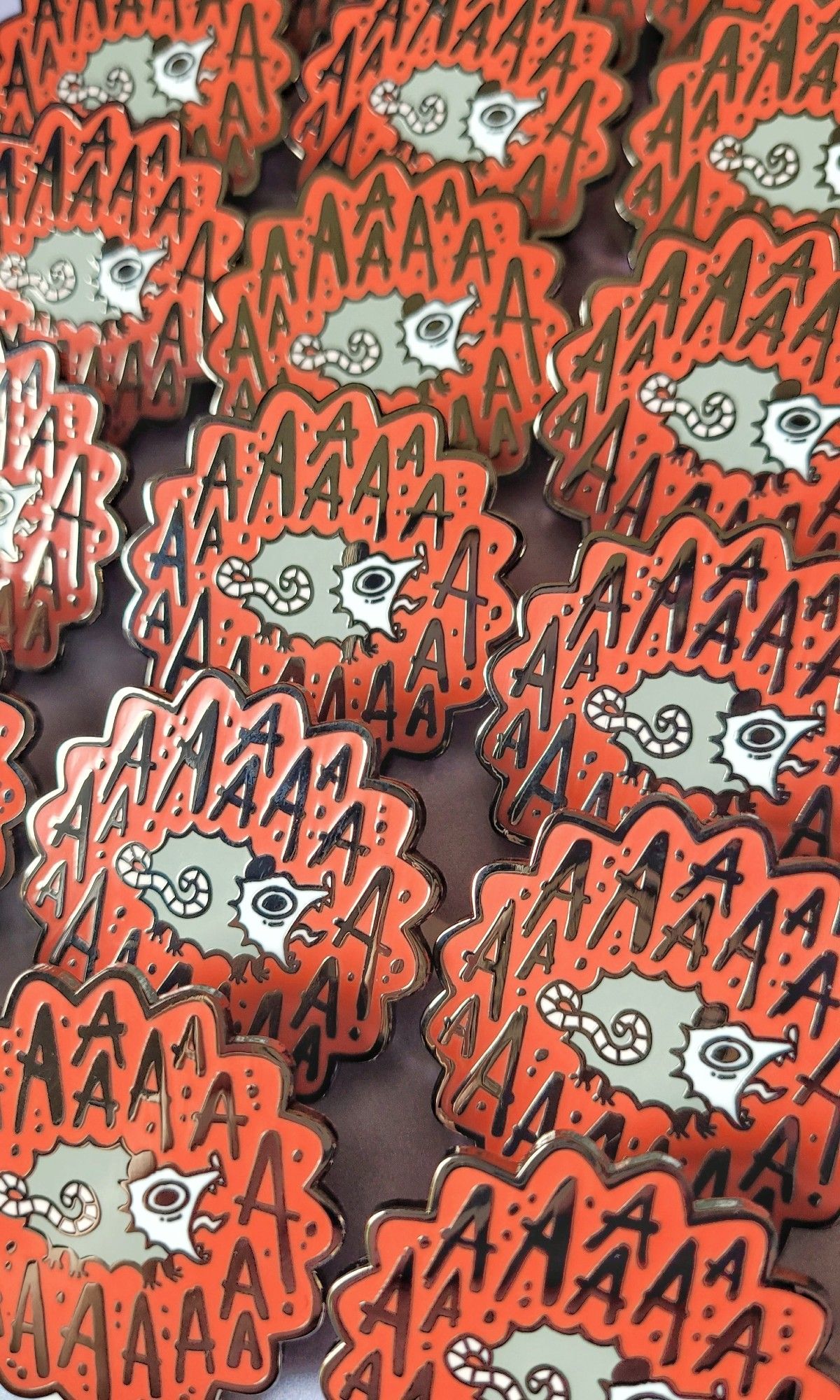 Photo of enamel pins of a red spikey speech bubble with a sceaming possum in the middle, surrounded by the letter A.