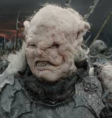 a rather pink and lumpy orc called Gothmog from the Lord of the Rings films wearing dark raggedy armor and grimacing. tufts of while hair on his head. one of his eyes is obscured partially by the lumpiness. 