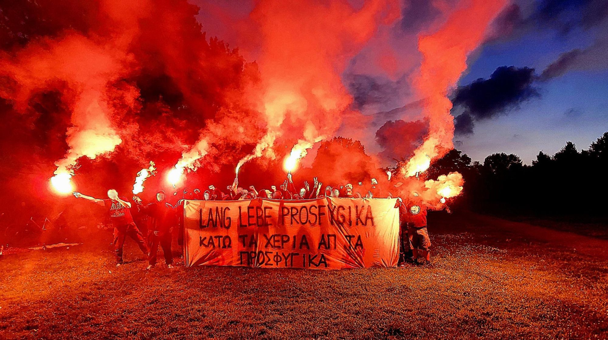 Ein Foto einer Gruppe mit Pyrotechnik und einem Transparent. Darauf steht "Lang lebe Prosfygika", darunter auf Griechisch "Hände weg von Prosfygika"