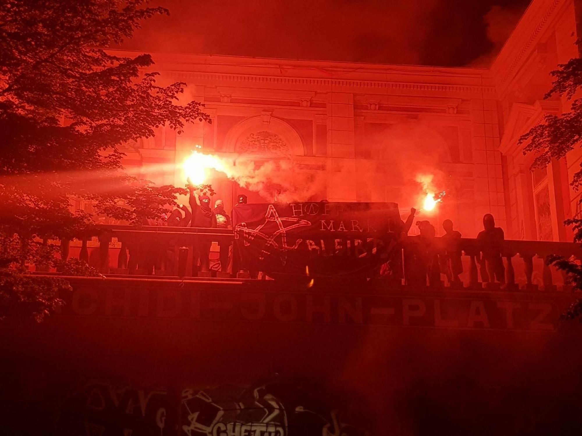 Auf dem Balkon der Roten Flora hält eine Gruppe Vermummter Bengalos und ein Transparent. Darauf steht "AZ Hafermarkt bleibt"