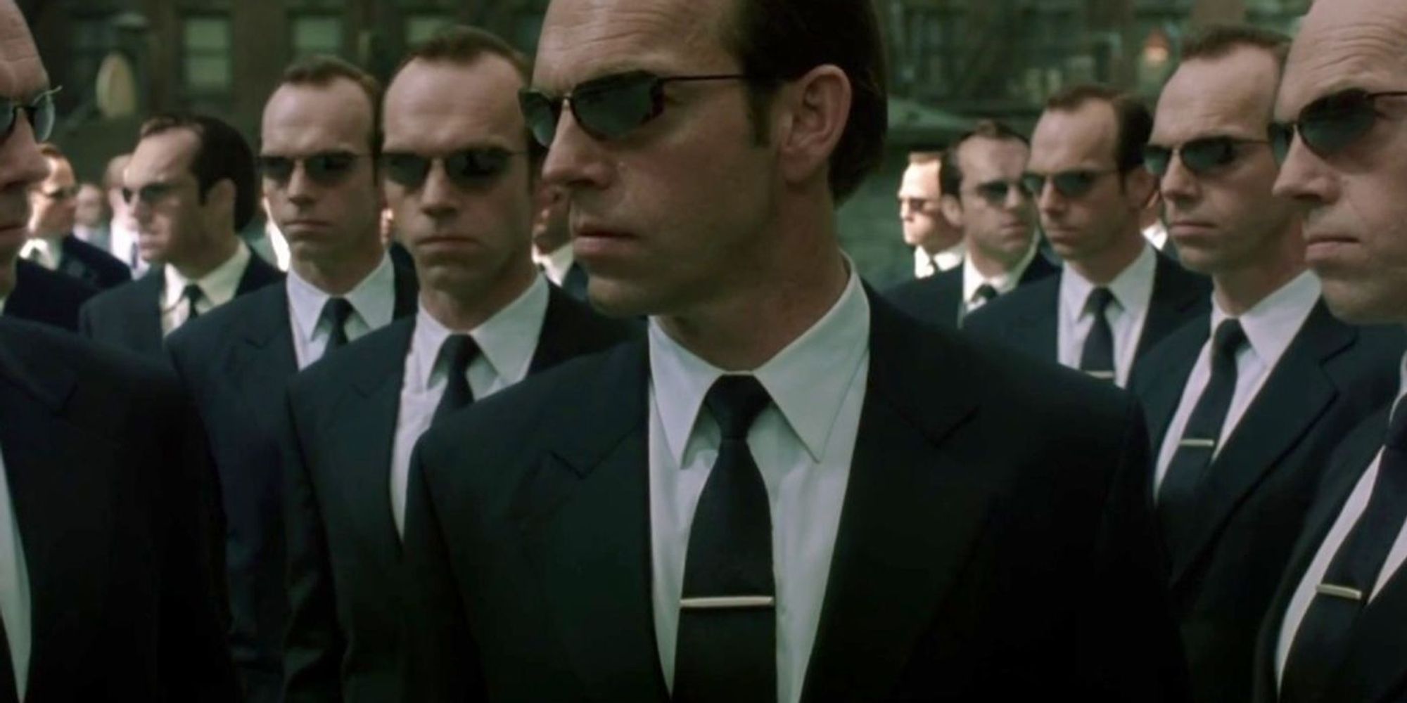 Das Bild zeigt eine Szene aus dem Film Matrix, in der mehrere Versionen von Agent Smith zu sehen sind. Agent Smith, gespielt von Hugo Weaving, trägt wie üblich einen schwarzen Anzug, ein weißes Hemd, eine schwarze Krawatte und eine Sonnenbrille. In dieser Szene sind viele identische Kopien von ihm zu sehen, was seine Fähigkeit symbolisiert, sich zu vervielfältigen und die Matrix zu kontrollieren. Die Szenerie wirkt bedrohlich und gleichförmig, da alle Agenten exakt gleich aussehen und dieselben Bewegungen und Mimik aufweisen, was eine unheimliche Atmosphäre erzeugt.