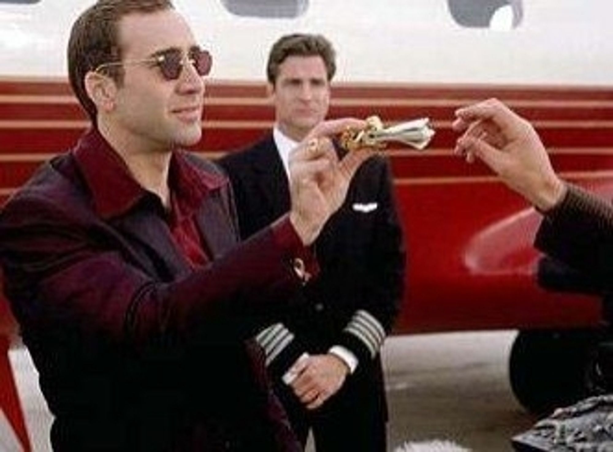 Das Bild zeigt eine Szene aus dem Film Face/Off (1997) mit Nicolas Cage in einer ikonischen Rolle. In der Szene trägt Cage eine Sonnenbrille und einen eleganten, dunkelroten Anzug. Er steht vor einem Privatjet und übergibt lässig Geld in einer Geldklammer an eine andere Person, wobei man seine entspannte und selbstbewusste Haltung erkennen kann. Im Hintergrund sieht man einen Piloten, der die Szene beobachtet. Dies ist eine Szene, in der Cage den kriminellen Charakter Castor Troy spielt, der für seine exzentrische Art und seinen dekadenten Lebensstil bekannt ist.