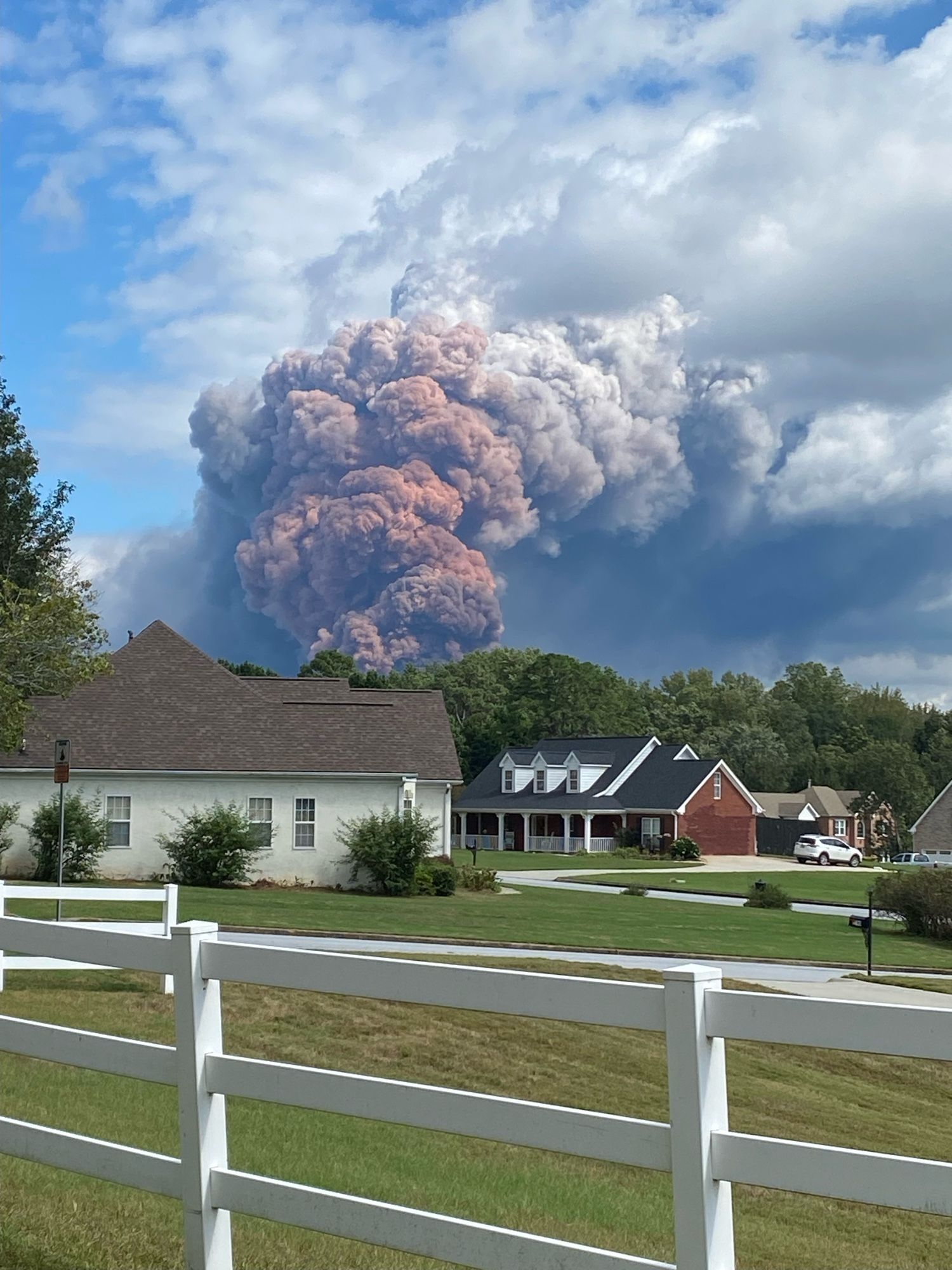 A suburban neighborhood in the middle of the day. Billowing in the sky above charming houses, green lawns, and white picket fences is a gargantuan plume of orange and gray smoke. It is apocalyptic, domineering, swarming the sky.