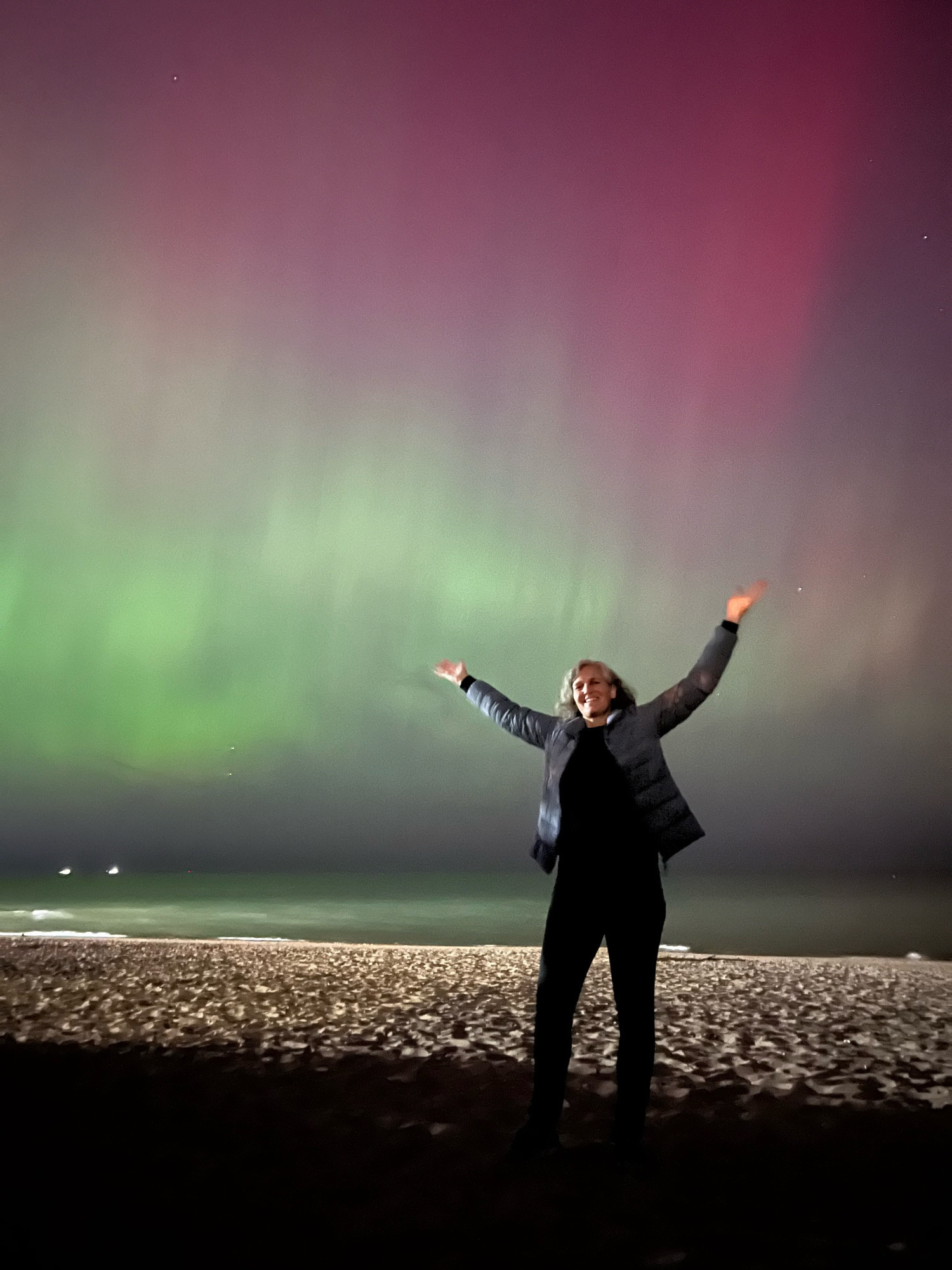 Photo of me standing on a beach at night with my arms up. Behind me, the sky is streaked with green and red pillars of aurora borealis.