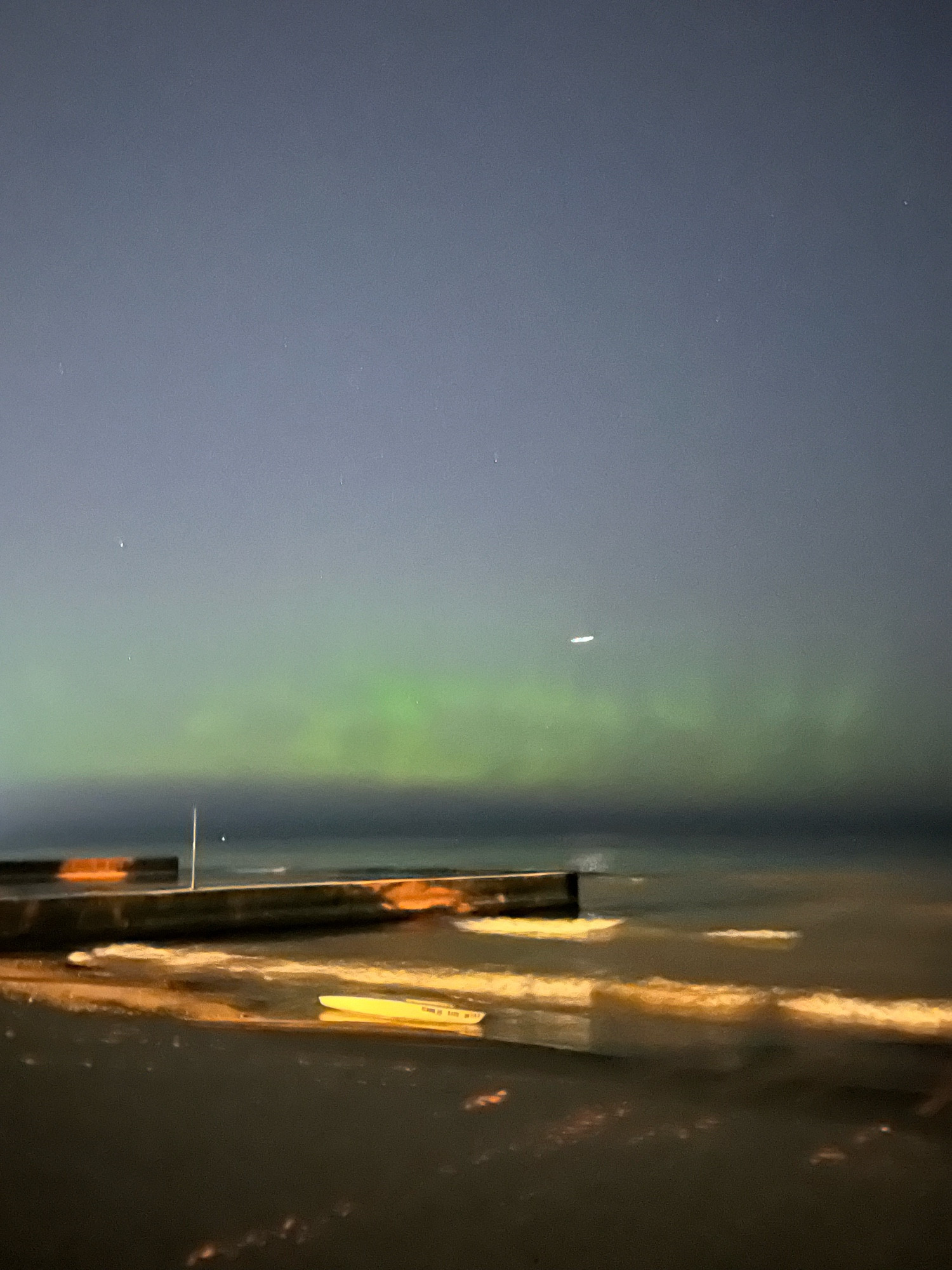 A beach at night with some waves and a pier. In the sky are faint pillars of green light from the aurora borealis