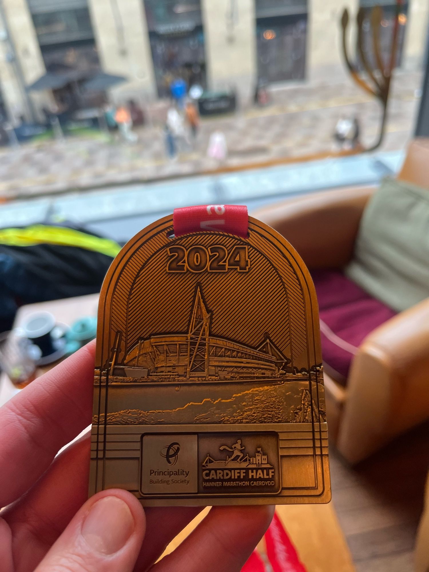 My hand holding up a Cardiff Half Marathon medal. The medal has ‘2024’ written on it with an engraving of the Millennium Stadium. It is copper coloured.