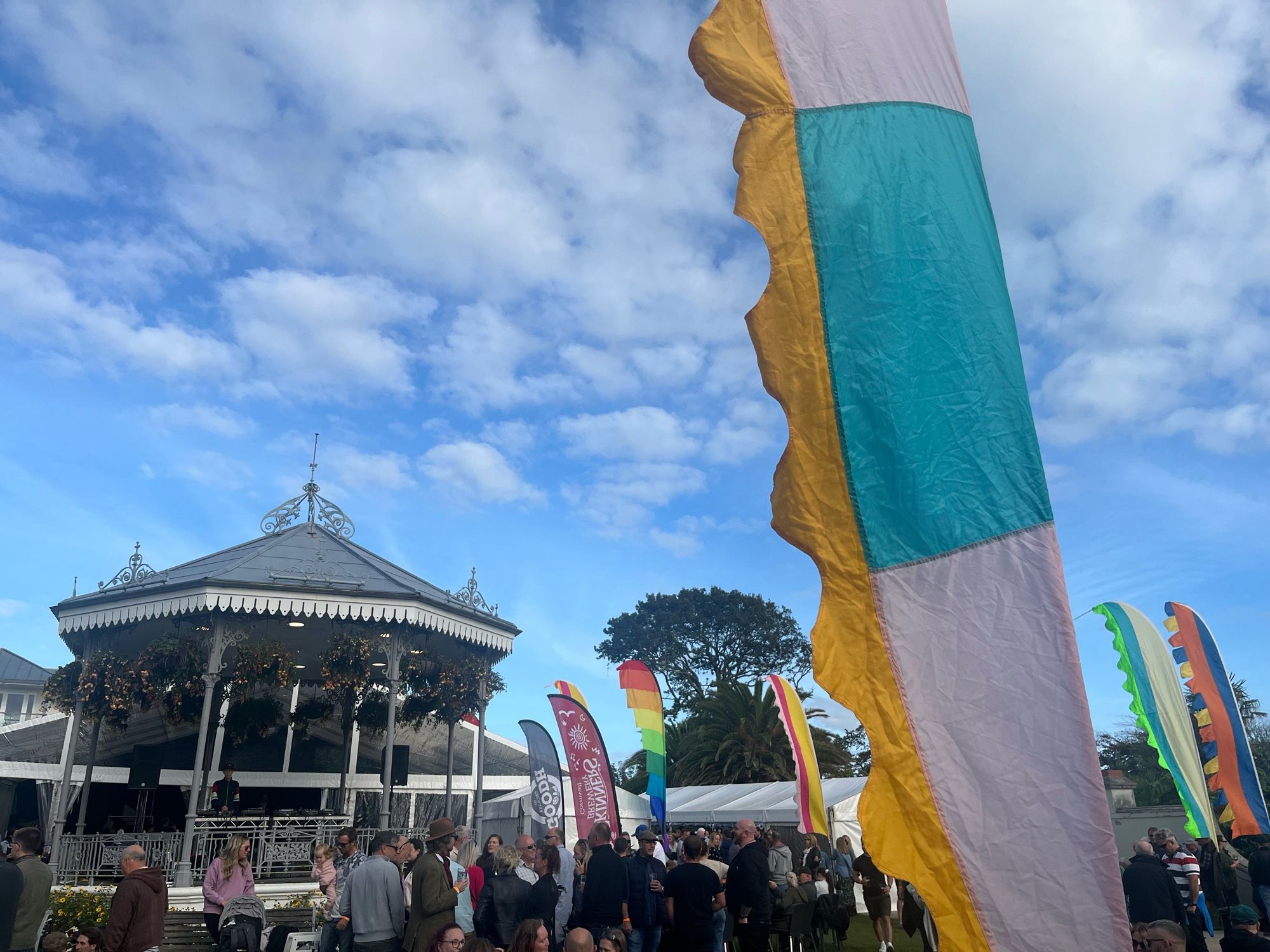 A flag blowing in the wind. In the background in an old fashioned bandstand. The sky is blue with fluffy clouds. People are milling around in front of the bandstand.