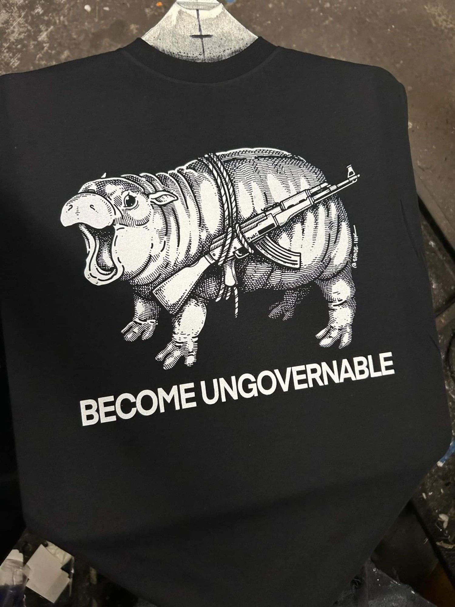 A black tee shirt featuring the baby hippo Moo Deng with a gun tied around its body with the caption "BECOME UNGOVERNABLE"