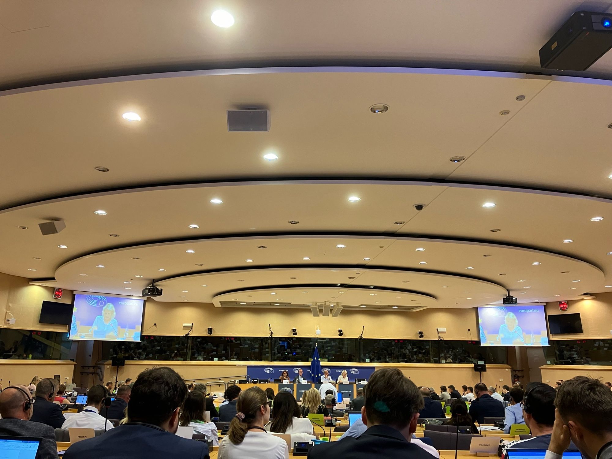 A bright circular committee room.
In the foreground, the backs of a crowd of seated people. In the centre, a barely visible stage with a blue background. Either side two large screens show the speaker.