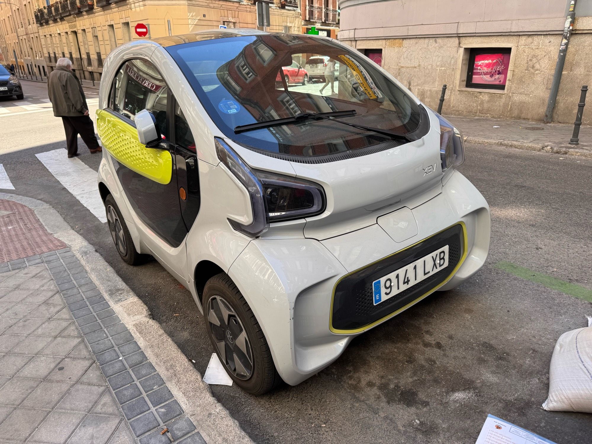 A street-parked XEV Yoyo, a teeny-tiny electric city car with quirky styling and a tiny footprint. It has a huge bubble windshield  and is primarily white with lime yellow accents.