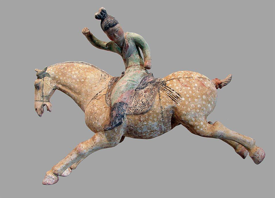 Ancient painted ceramic sculpture of a rider in traditional attire on a galloping spotted horse with raised front legs.