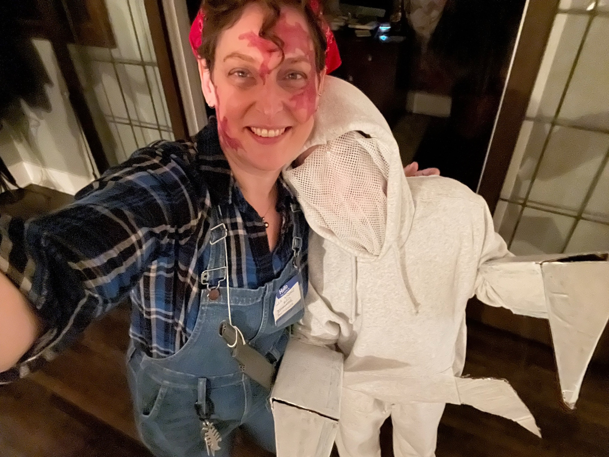 Me (white femme) as Stella (birthmarked bootlegger from Appalachian holler) and kid as Ghostdaddy (mantid godmonster). I’ve got noticeable sprawling facial birthmarks, a blue plaid shirt, overalls, a rifle, and a name tag. Kid has white sweats, a mesh face covering, an extra pair of legs thru the sweatshirt pocket, and cardboard mantid arms.