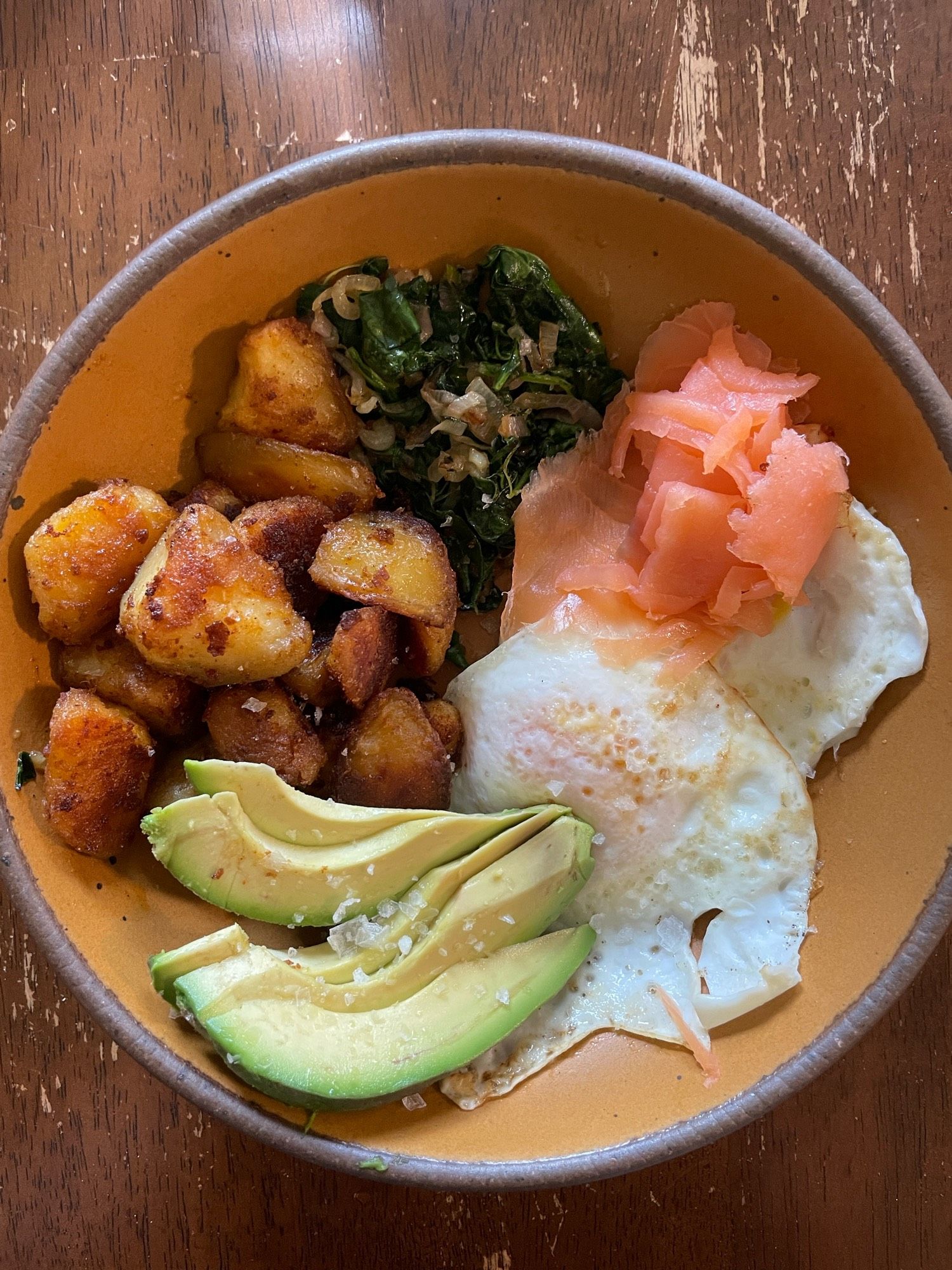 an orange breakfast bowl consisting of two fried eggs, a quarter avocado, roasted potatoes, garlic/shalloy kale and some smoked salmon