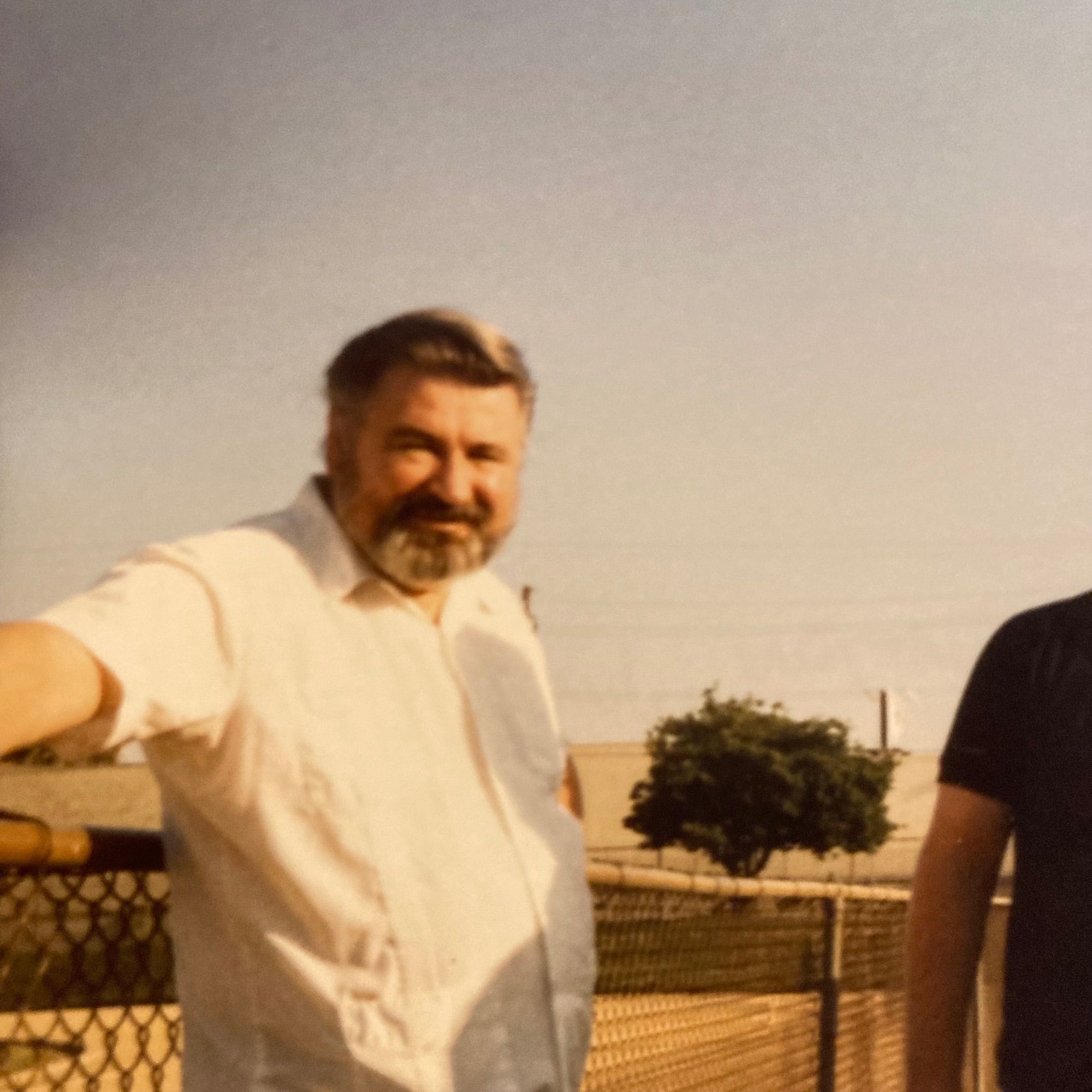 A photograph of a man, taken in the 1980s. He has black, greying hair and a beard, and a collared shirt on. He is leaning casually against a chain-link fence.