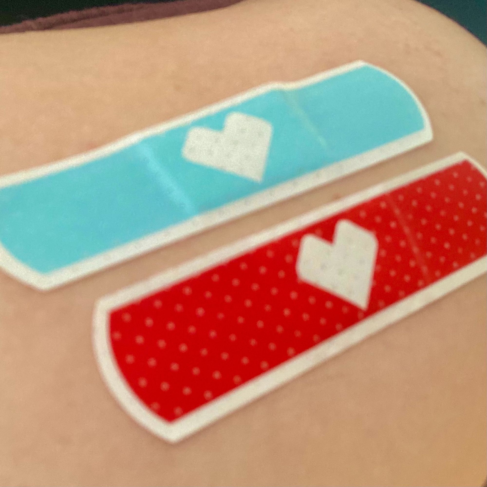 A photograph of two bandaids - one cyan, the other red - on an upper arm