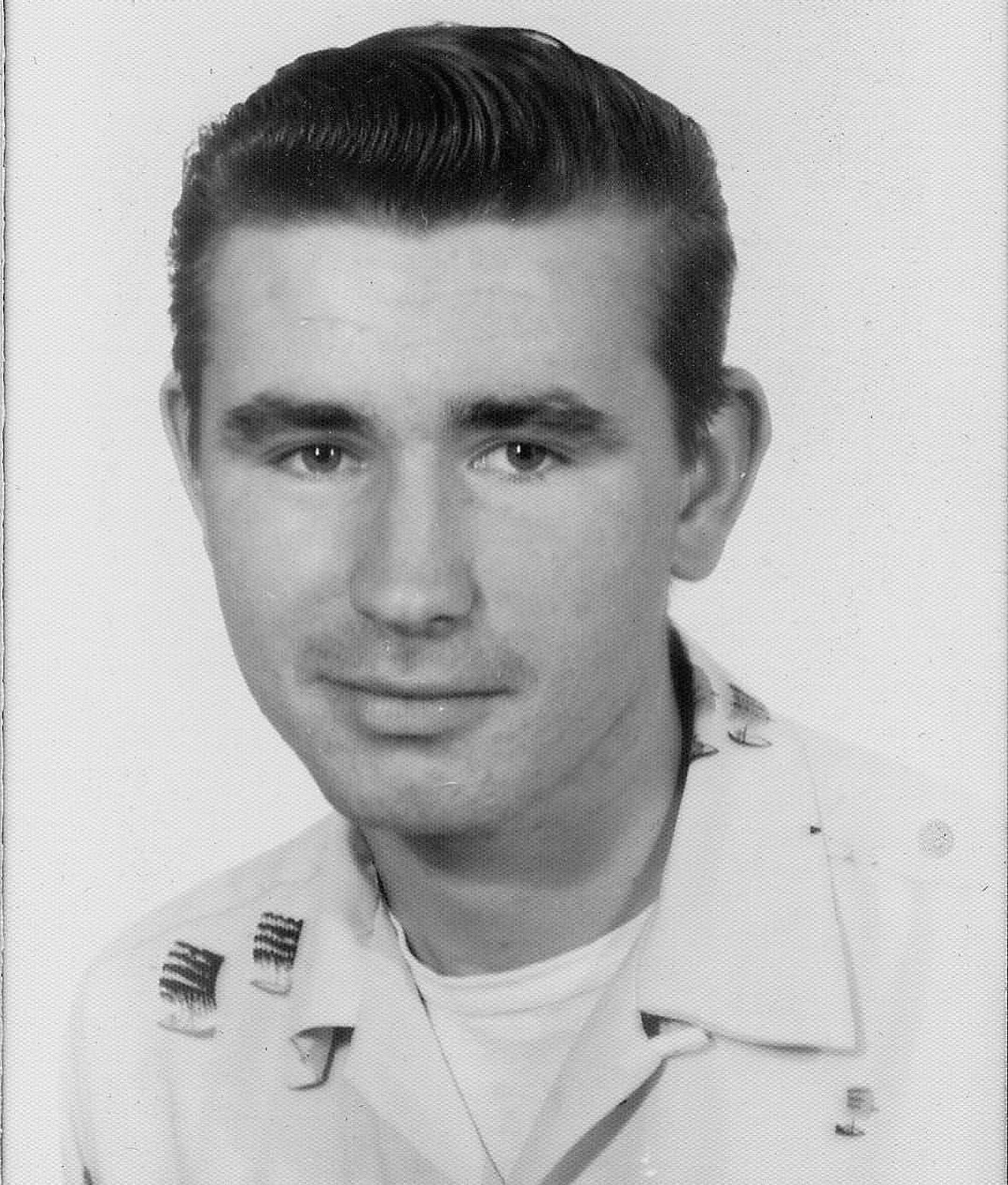 A black and white photograph of a young man, taken in the 1950s. He has black, slicked back hair, and a patterned collared shirt over a t-shirt on.