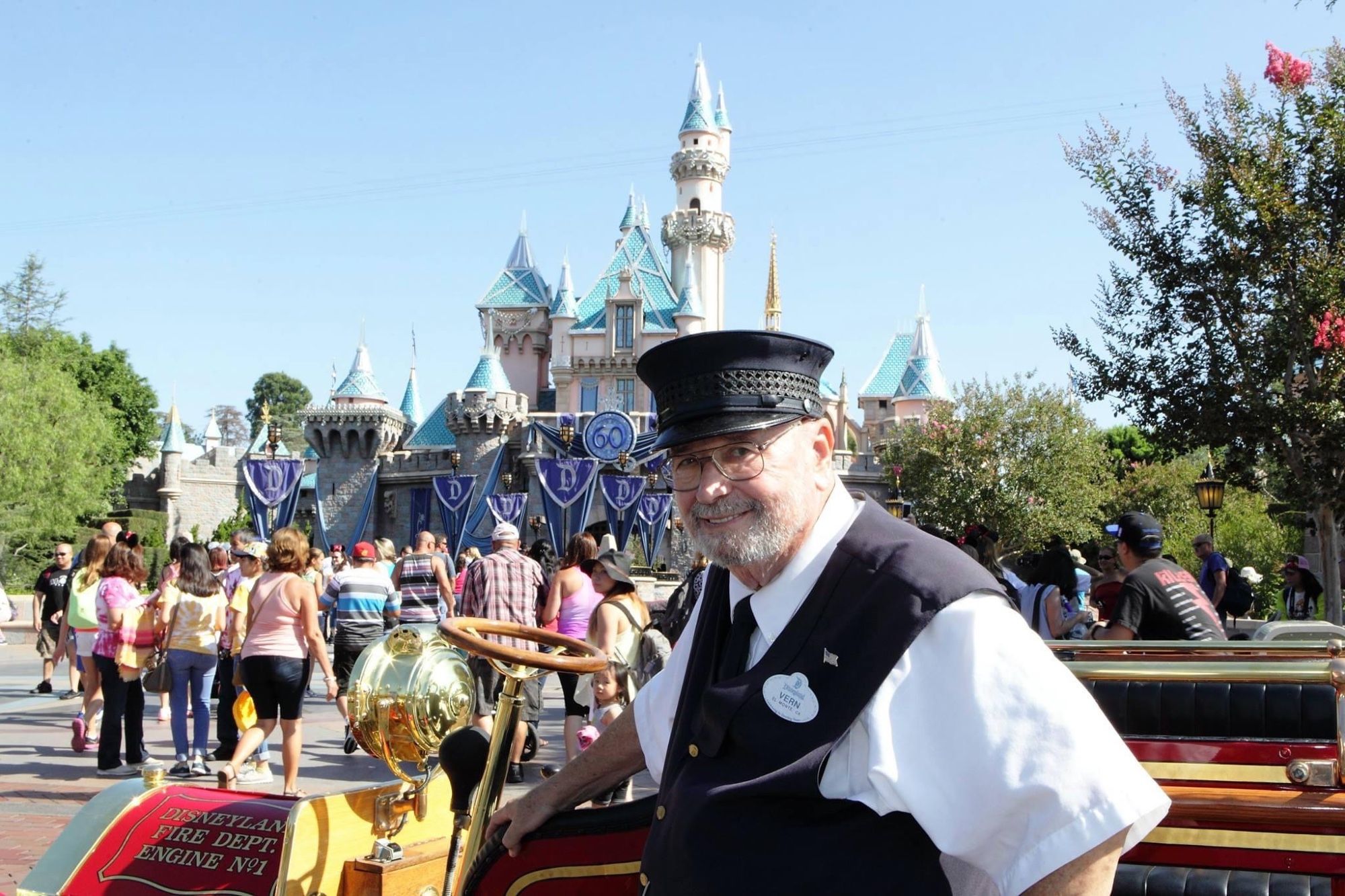 A photograph of a man, taken in the 2015. He has a grey beard, and is wearing a Fireman's cast member uniform. He is leaning against the Disneyland firetruck, and is in front of Sleep Beauty Castle at Disneyland. The Castle is decorated for Disneyland's 60th anniversary.