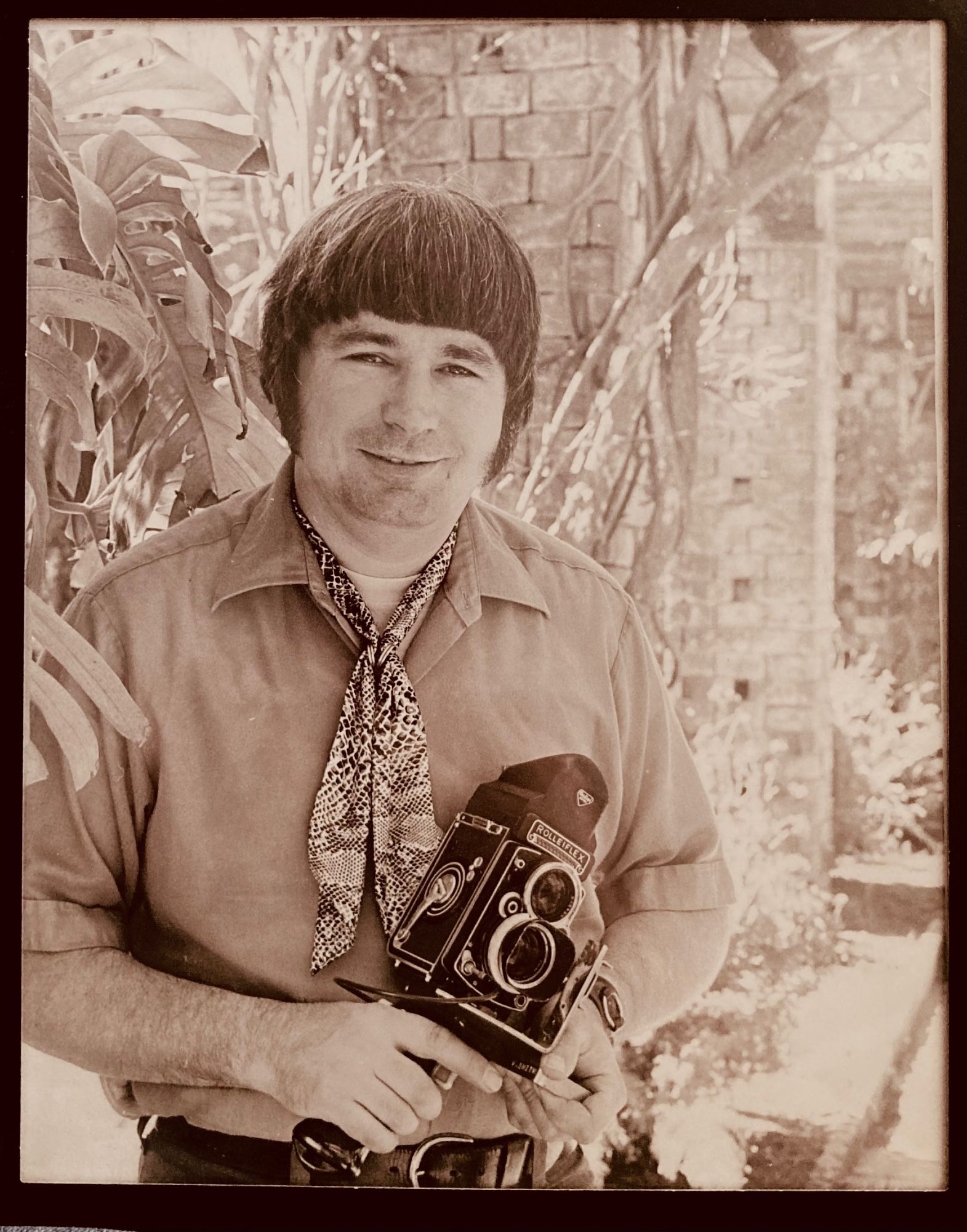 A sepiatone photograph of a man, taken in the 1970s. He has black, "Beatles" style hair, and a patterned cravat over a short sleeved, collared shirt. He is holding a Rolleiflex camera on a pistol grip.