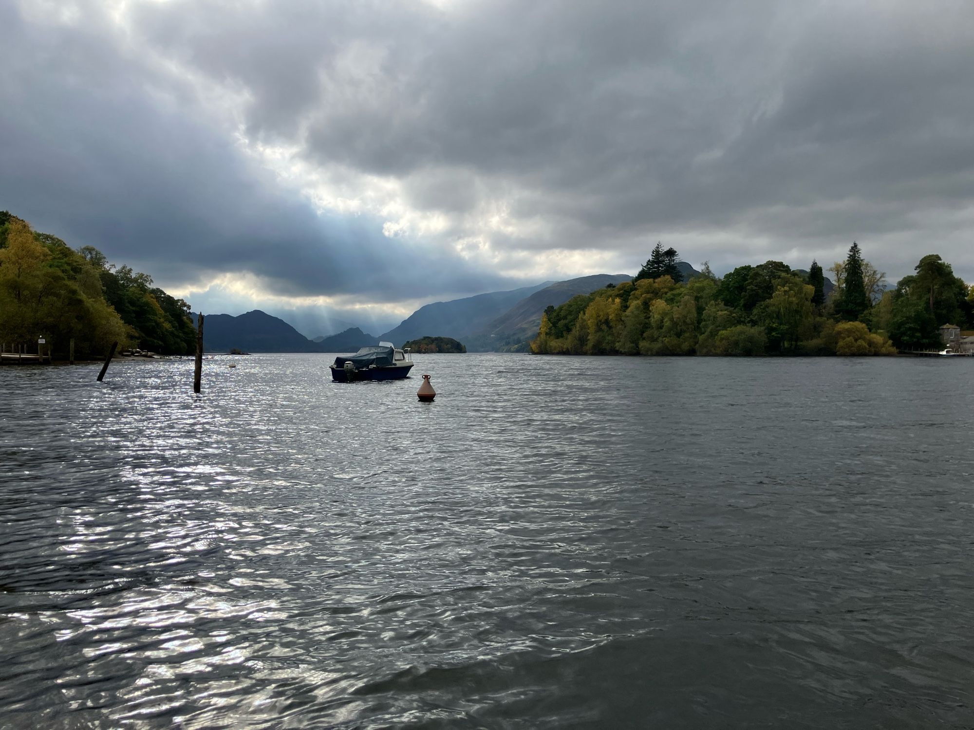 Ein See. Der Himmel ist bewölkt, die Sonne bricht durch die Wolken und beleuchtet Teile der Berge im Hintergrund. Auf dem See ankert ein Boot und es ist eine bewaldete kleine Insel zu sehen.