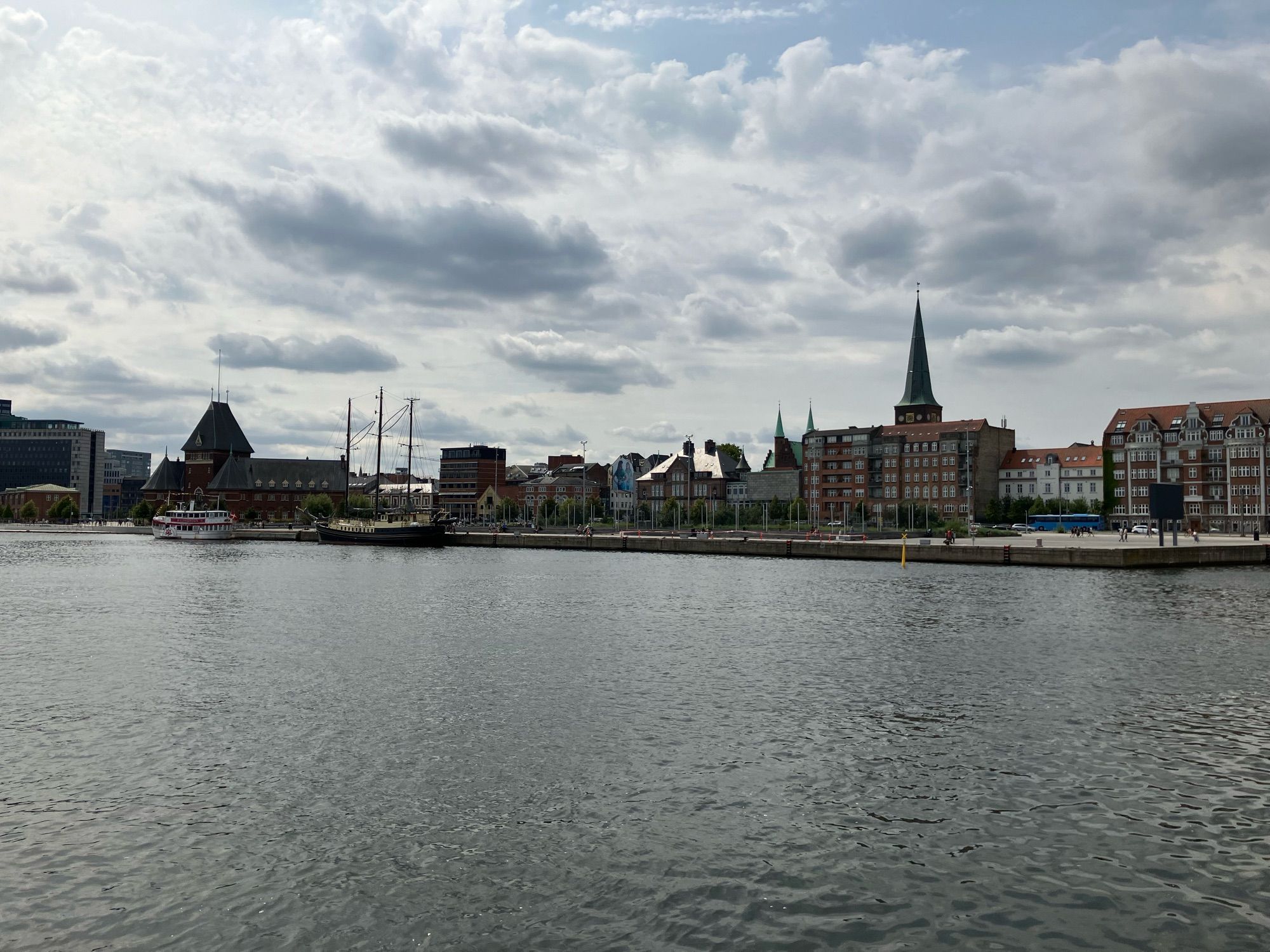 Blick von Wasser auf Aarhus. Der Dom überragt die Gebäude, die dichter am Wasser stehen. Am Kai liegen ein Ausflugsdampfer und ein Segelschiff.