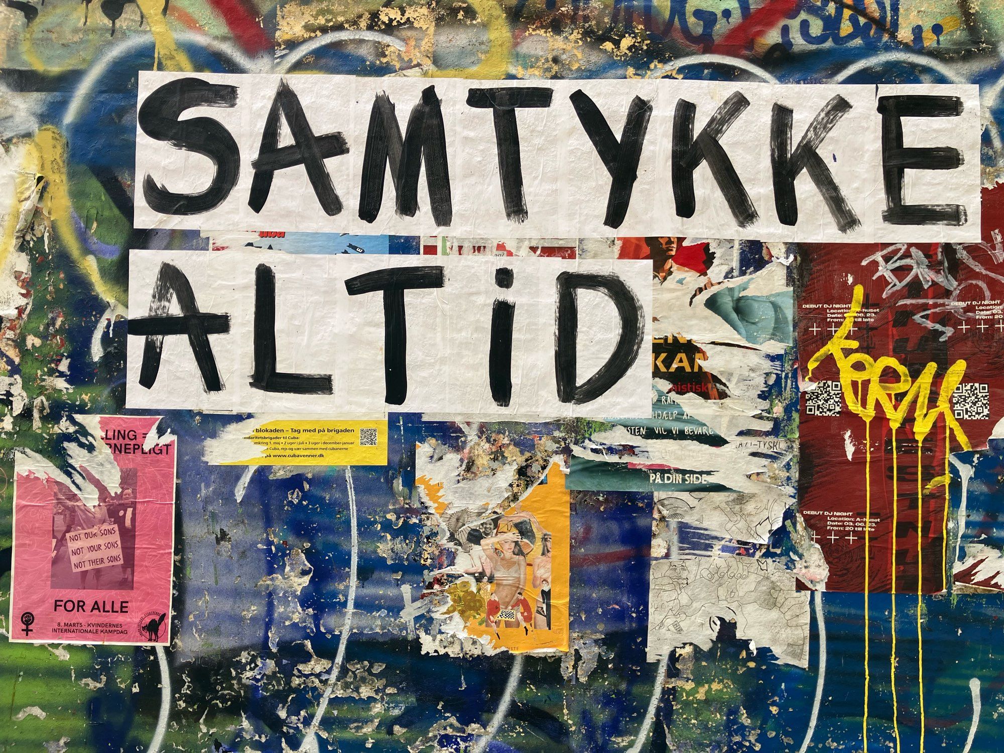 Eine Wand mit Graffiti und aufgeklebten Postern. Am klarsten zu erkennen ist der Schriftzug “samtykke altid” in schwarzen buchstaben auf weißem grund (laut online Übersetzer: “Immer Konsens / always consent”).