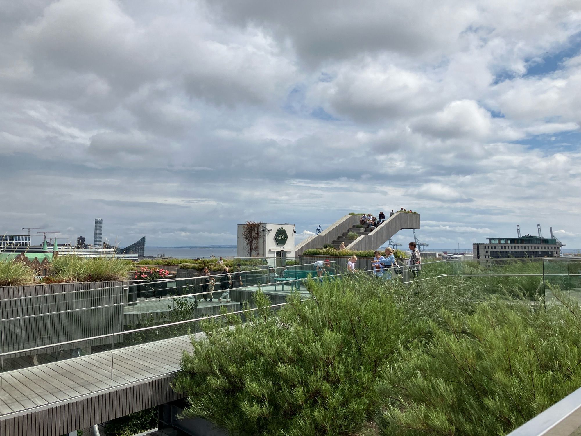 Blick über eine hölzerne Dachterasse mit Bergrünung und einer Treppe zu einer noch höheren Aussichtsplattform. Dahinter Aussicht auf Aarhus Ø, den alten Hafen und die Ostsee.