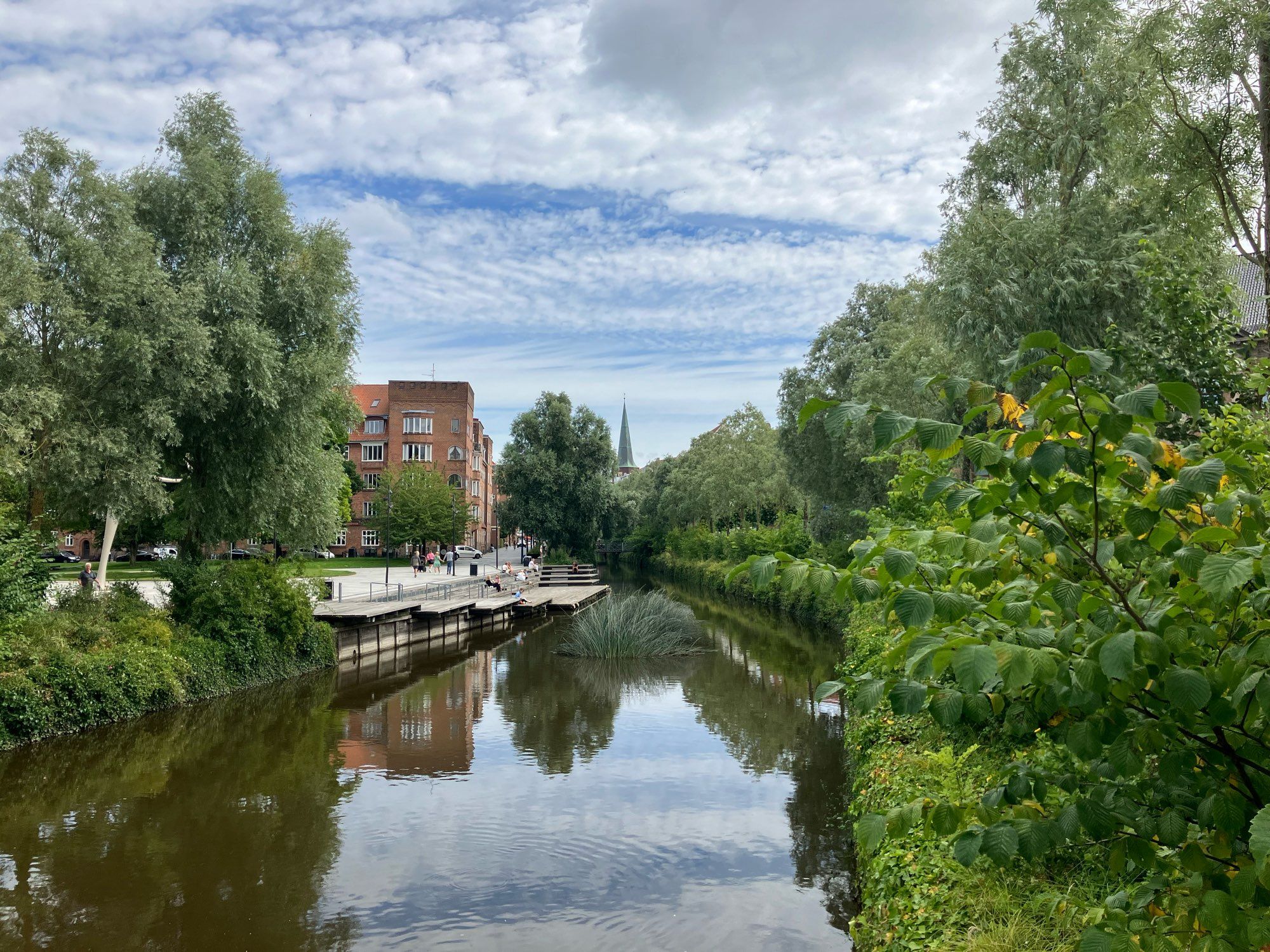 Blick von einer Brücke auf den Fluss in Aarhus. Das rechte Ufer ist komplett begrünt. Am linken Ufer ist die Begrünung durchbrochen durch hölzerne Plattformen zum sitzen. Die Bäume und ein Gebäude links des Flusses spiegeln sich im Wasser. In der Ferne ist ein Kirchturm zu sehen.