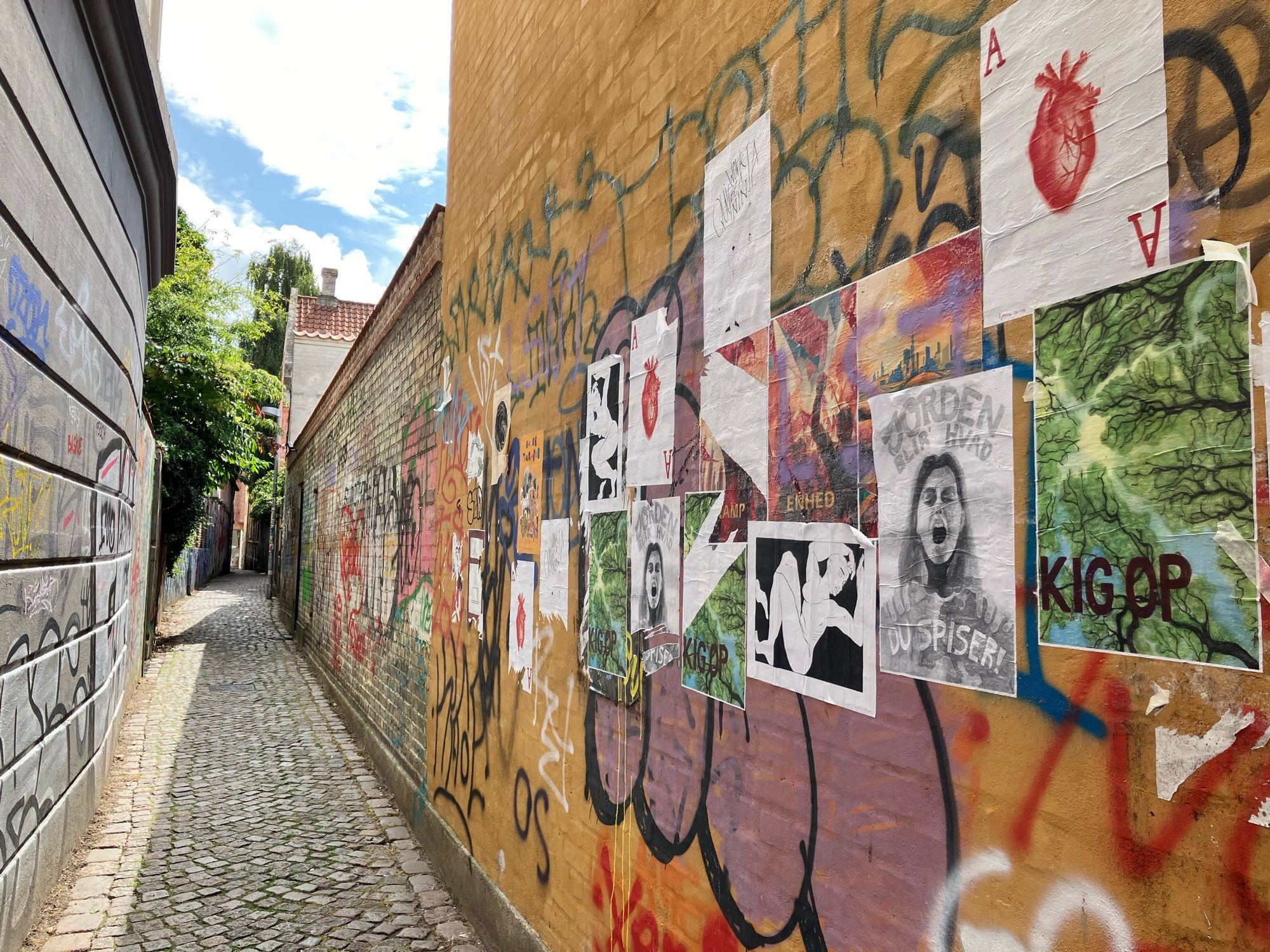 Eine schmale Gasse. Die Wände sind mit Graffiti besprüht, rechts hängen diverse Poster, einige schon teils abgerissen.