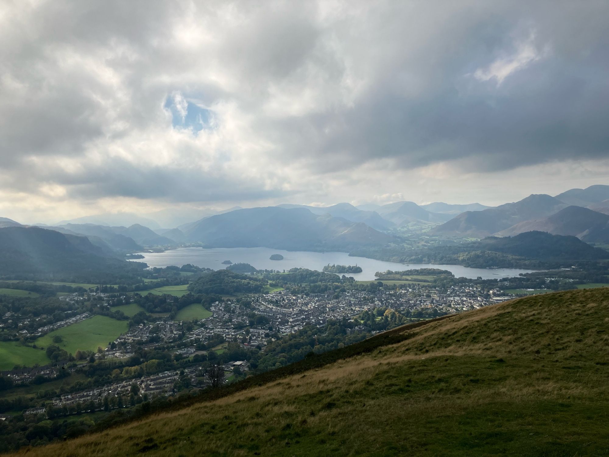 Blick von einem Berg auf den Ort Keswick. Weiße und graue Häuser auf einer Ebene, darum Wiesen und Bäume. Hinter dem Ort ein See und Berge. Die Sonne bricht durch die Wolken.