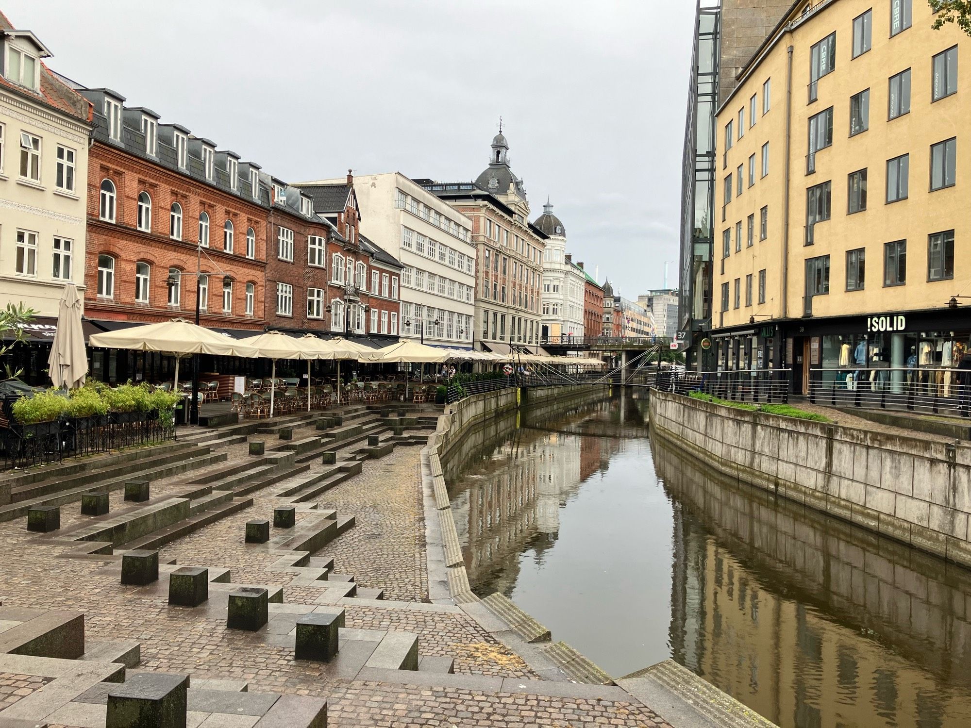 Blick von einer Brücke auf den Fluss in Aarhus. Links und rechts des Flusses stehen Gebäude, die sich im Wasser spiegeln.