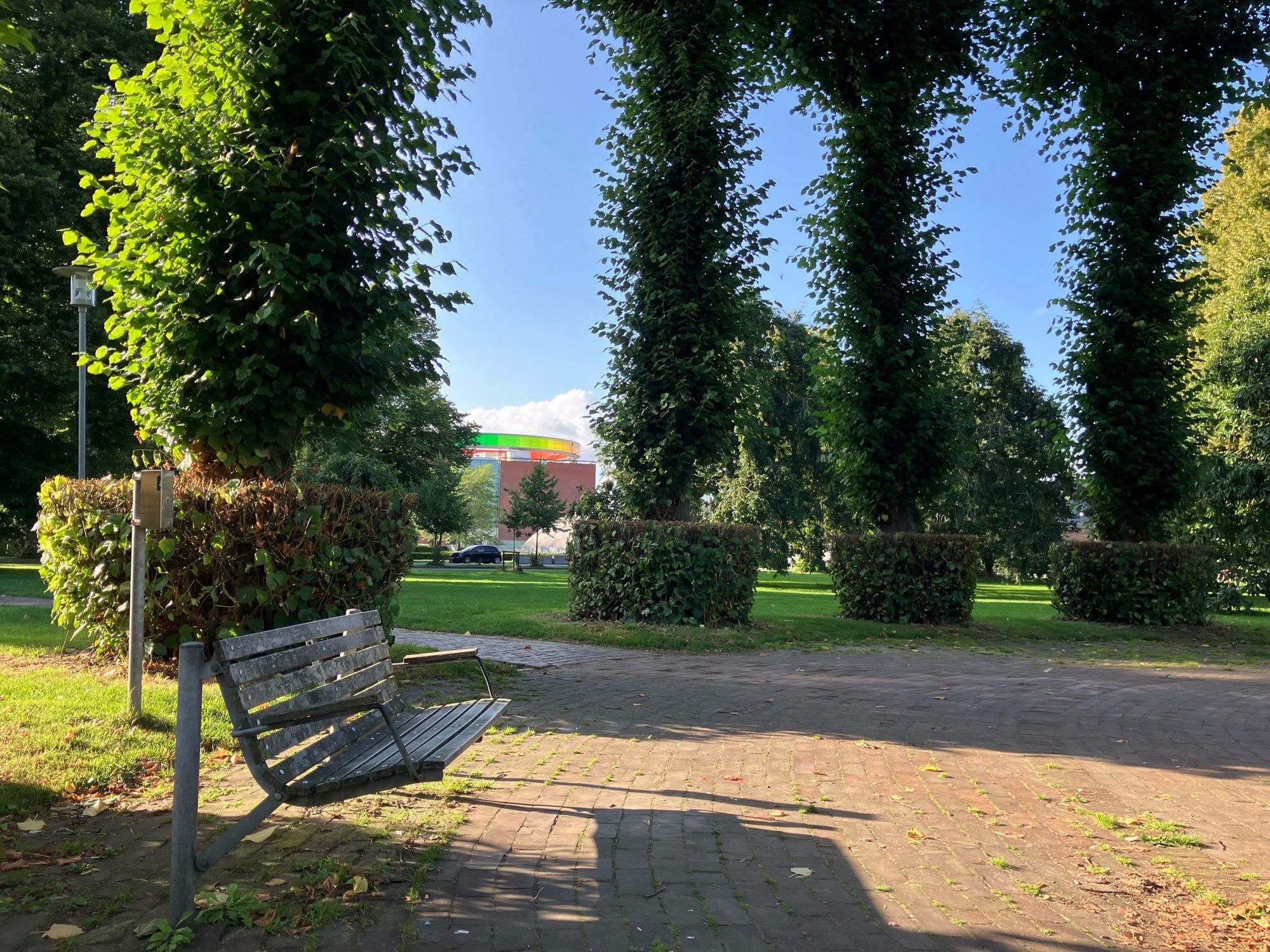 Eine Parkpank und Bäume in der Abendsonne. Zwischen den Bäumen hindurch sieht man das Aros Museum mir einem Regenbogen-farbenen Gang auf dem Dach.