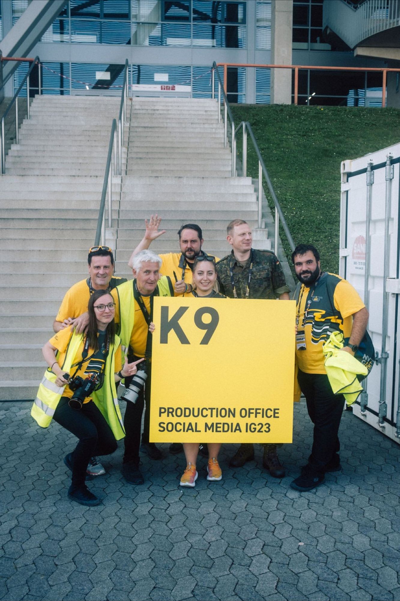 A group picture of the social media team at the Invictus Games Düsseldorf 2023. A group of people in uniform wave into the camera, as one holds a sign that reads: "(Entrance) K9: Production Office Social Media IG23