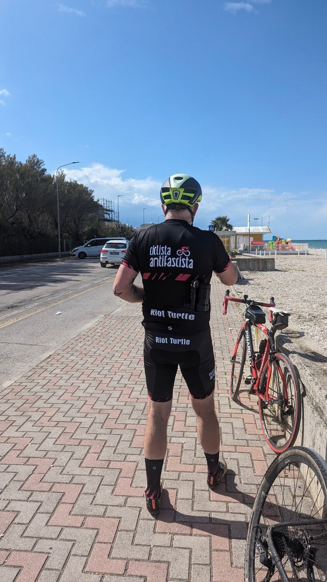 In der Bildmitte steht Riot Turtle im Ciclista Antifascista mit dem Rücken zur Kamera. Rechts von im steht sein Schwarze-rotes Pinarello Rennrad. Rechts im Bild ist auch ein Strand und das Meer zu sehen. Links ist der Küstenstraße und oben ein blauer Himmel.