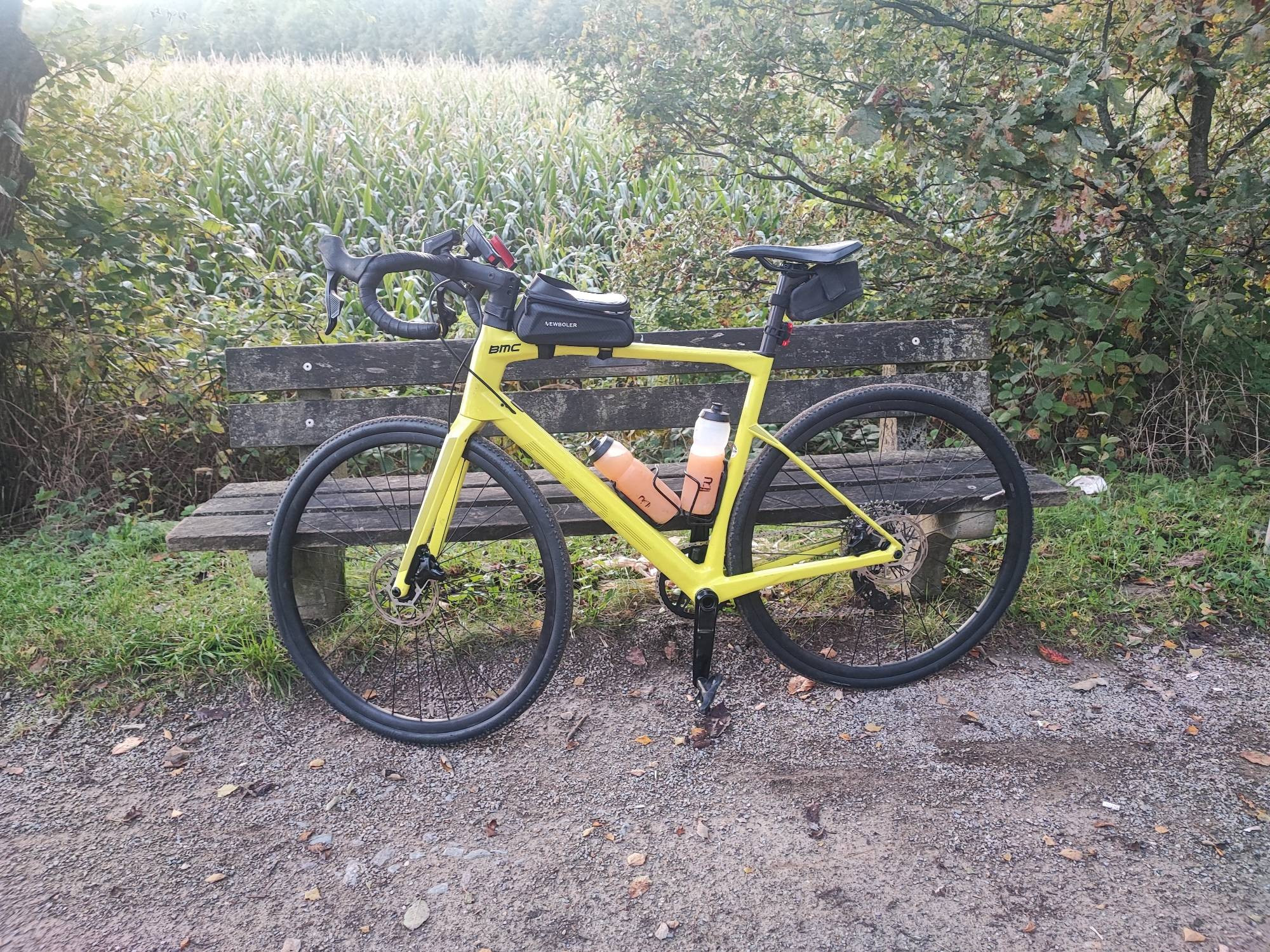 Mein Gelbes BMC Gravelbike steht gegen ein Bänkchen auf der Glückauftrasse zwischen Niedersprockhövel und Schee. Hinter das Bänkchen ist ein Maisfeld zu sehen. Vor dem Gravelbike der Gravelradweg Glückauftrasse.