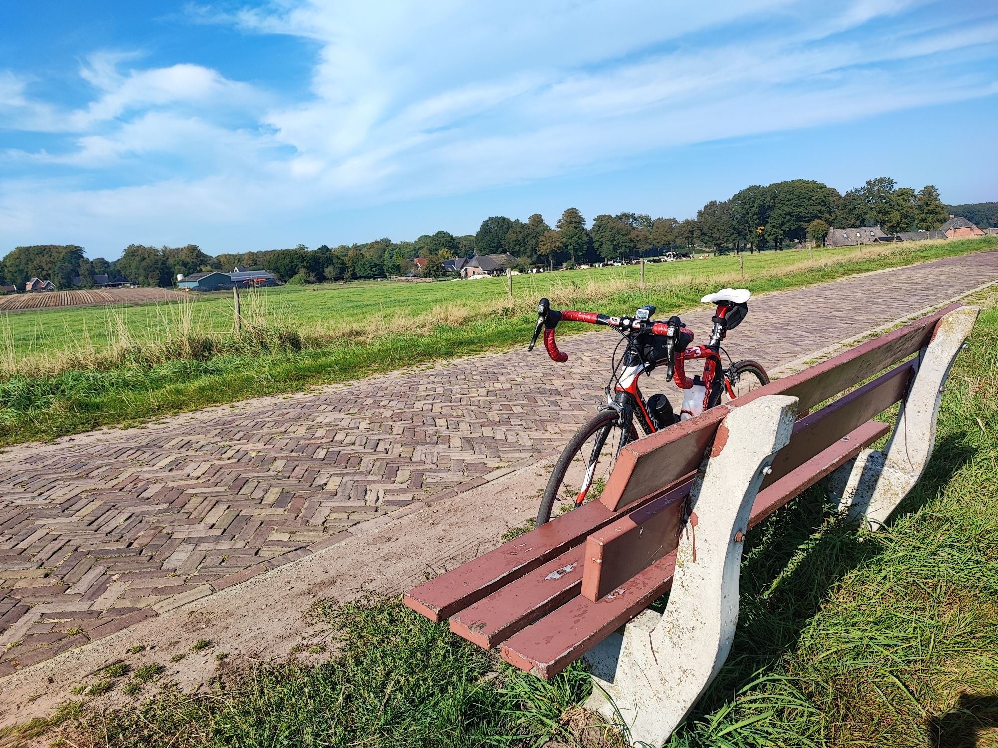 Mein Schwarze-rotes Pinarello Rennrad steht neben eine Straße mit Kopfsteinpflaster gegen ein Bänkchen. An beiden Seiten der Straße sind Wiesen zu sehen. Im Hintergrund mehrere Bauernhöfe und Bäume.