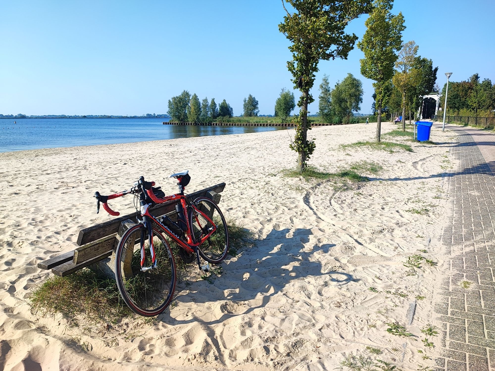 Mein Schwarze-rotes Pinarello Rennrad gegen die Rückseite einer Bank auf einem Strand an einer Seeplatte. Links ist der See zu sehen, rechts der Radweg.