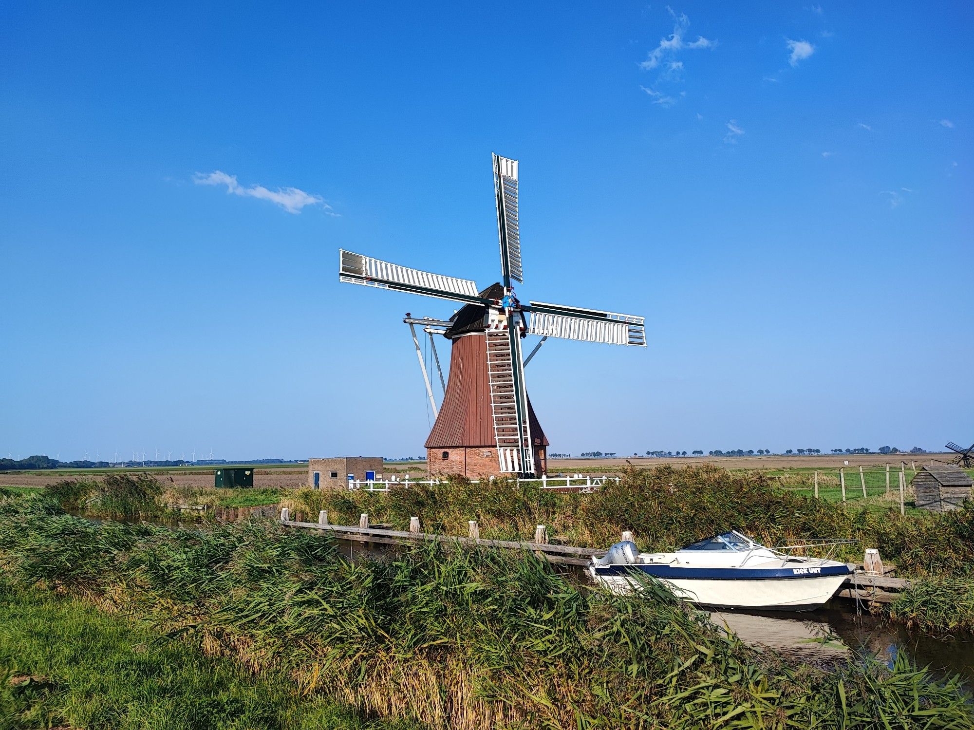 Im Vordergrund ist eine Wiese und Riet entlang ein Kanal zu sehen. Im Kanal ist auch ein weißes Boot zu sehen. In der Mitte vom Bild steht ein alter traditionelle niederländische Windmühle.