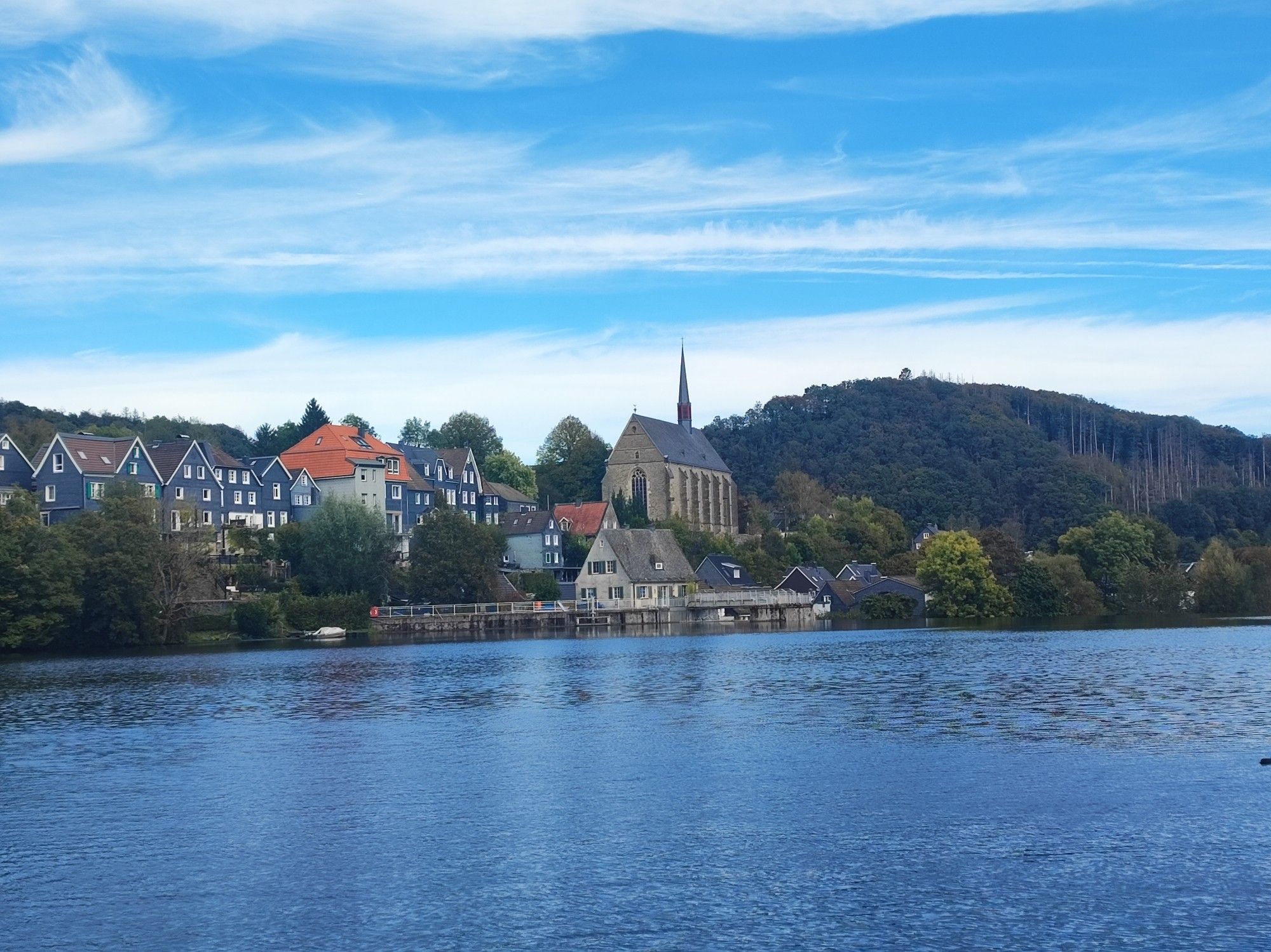 Im Vordergrund ist der Beyenburger Stausee zu sehen. Am Ufer die Kirche und einige Häuser im Ortskern von Wuppertal Beyenburg. Rechts hinten ist ein bewaldeten Hügel zu sehen.