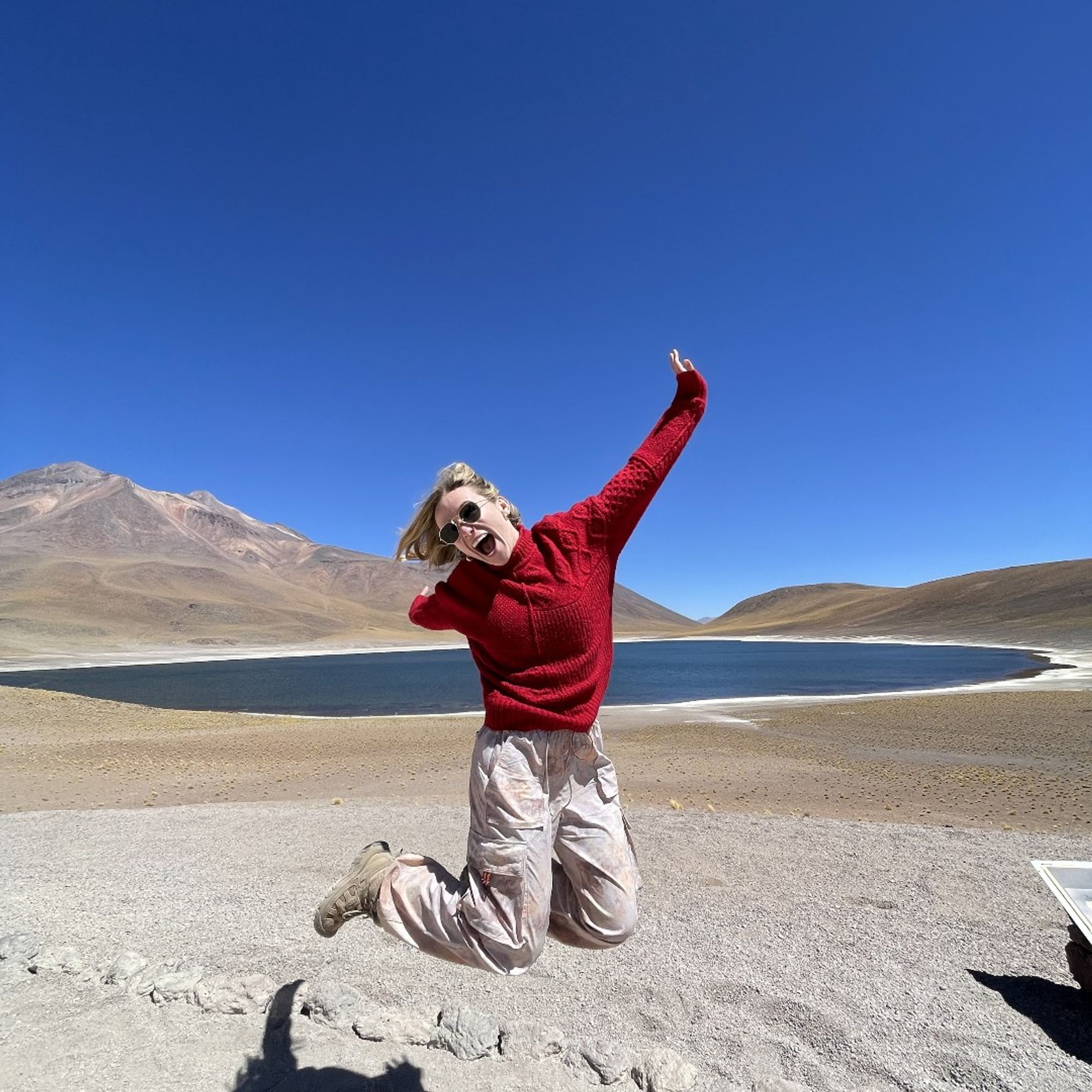 Purdue Veterinary Student Allison Jones in a red sweater leaping mid-air with a scenic backdrop of a mountain and a serene blue lake under a clear sky in Chile.