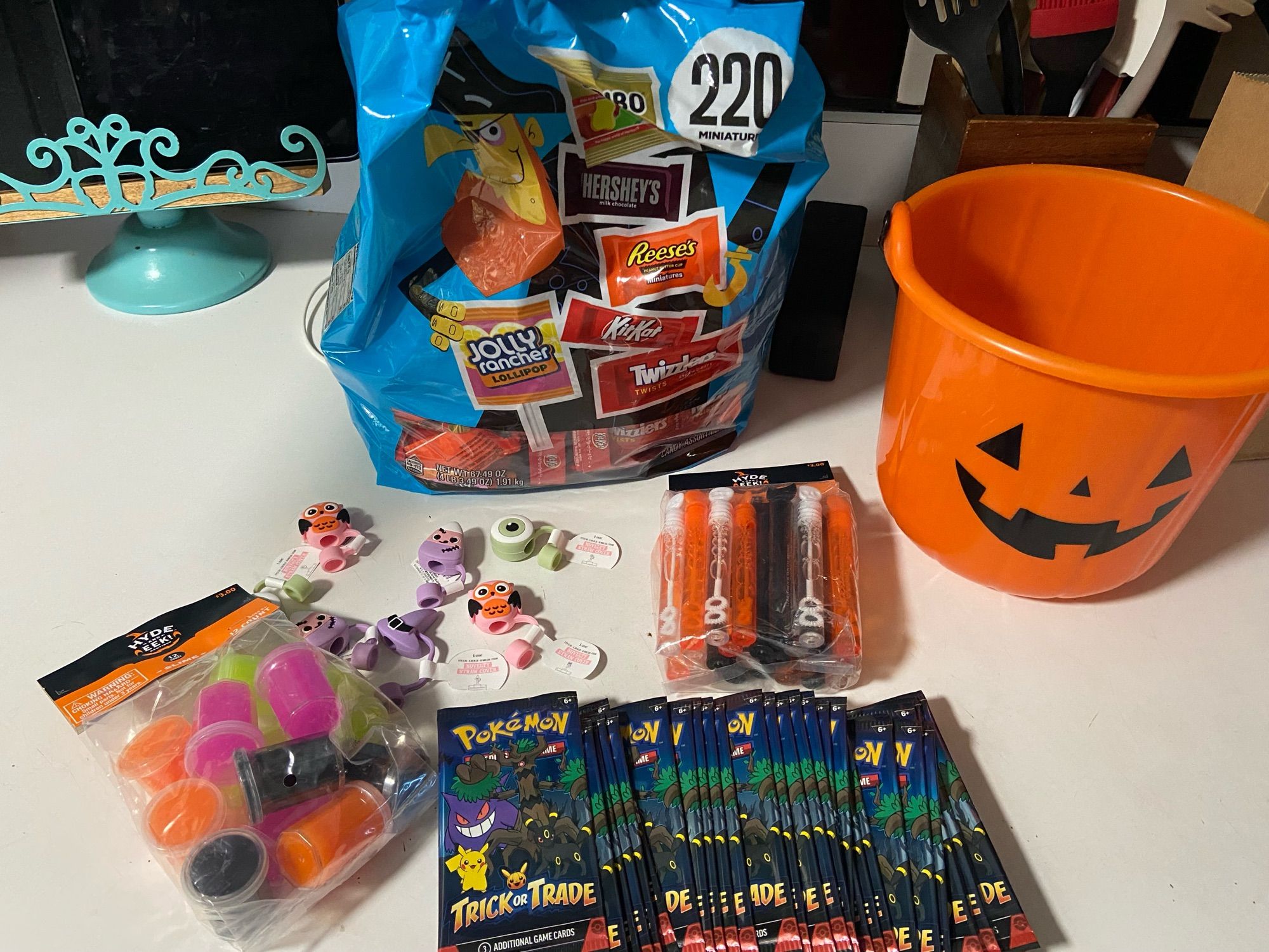 A picture of items to hand out for trick or treaters, including a bag of assorted candy (gummy bears, hershey’s, Reese’s, Kit Kat, Twizzlers, and jolly ranchers), tubs of Halloween colored slime, small tubes of bubbles, and small packs of Pokémon cards.