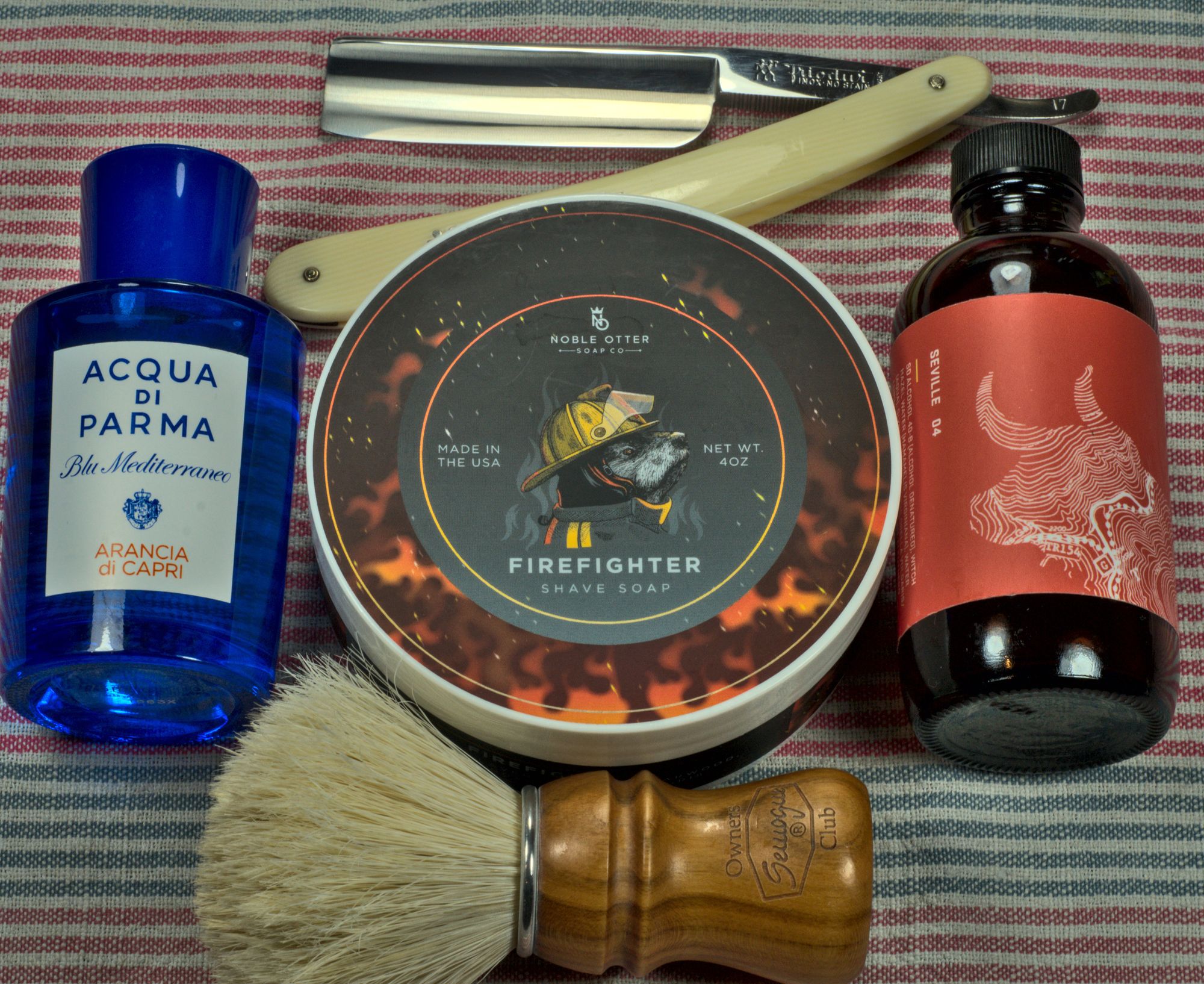 A tub of NO Firefighter soap between bottles of BaM Seville splash and AdP Blu Mediterraneo Arancia di Capri frag, a SOC boar brush, and a chonky Frio 17
