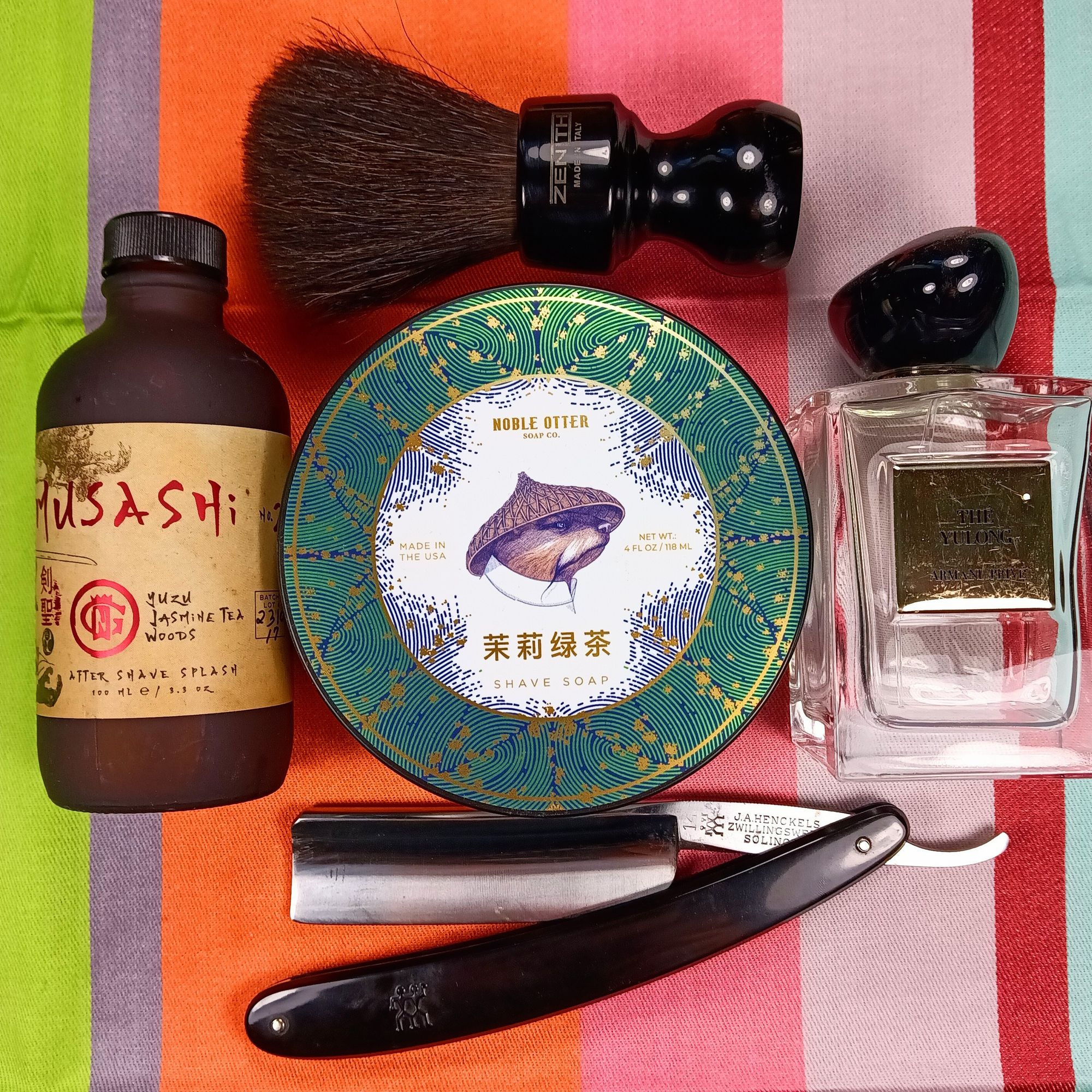 A tub of NO 茉莉綠茶 soap surrounded by bottles of GN Musahi spash, GA Thé Yulong frag, a pretty Zenith Horse brush, and a big Henckels 14 razor.
