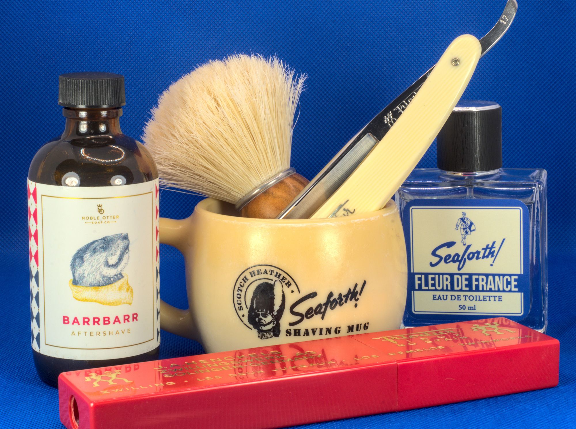 A chonky Frio 17 and a SOC boar brush in a vintage Seaforth! shaving mug between bottles of NO Barrbarr splash and Seaforth! Fleur de France frag with a bright red Friodur razor case in  the foreground