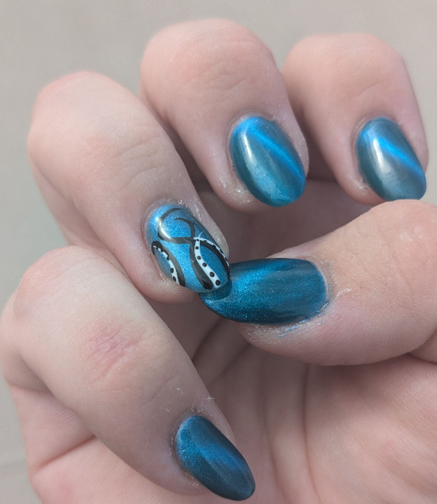 A photo of a hand with long oval nails painted with a teal magnetic polish, the ring fingernail has black and white tentacles