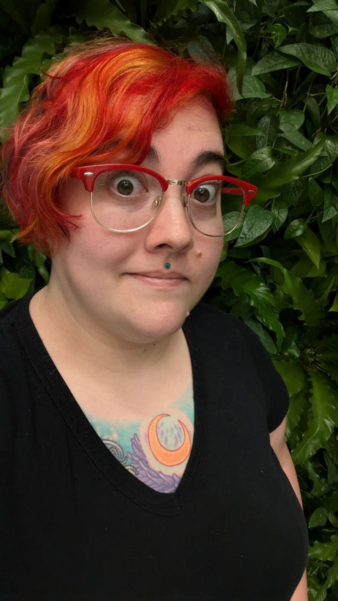 a selfie of Tom with red and orange hair, standing in front of a lush leafy green (faux) foliage wall