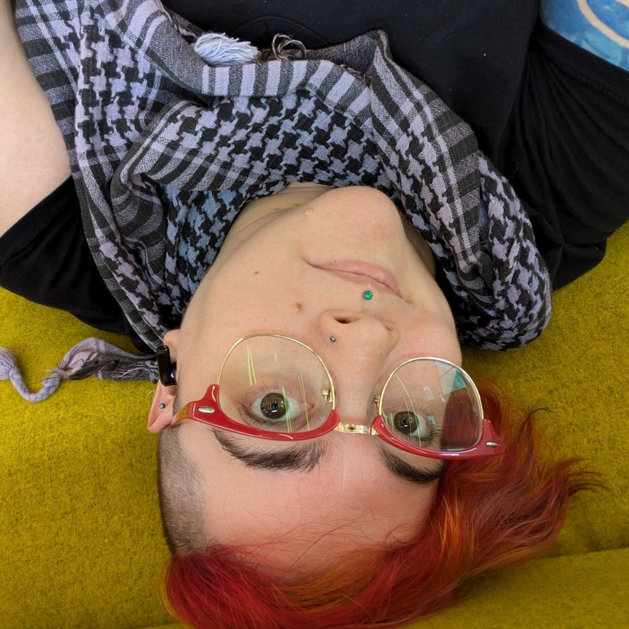 a selfie of Tom taken upside down, Tom has fiery red hair, red glasses, a blue and black keffiyeh, and a black t-shirt