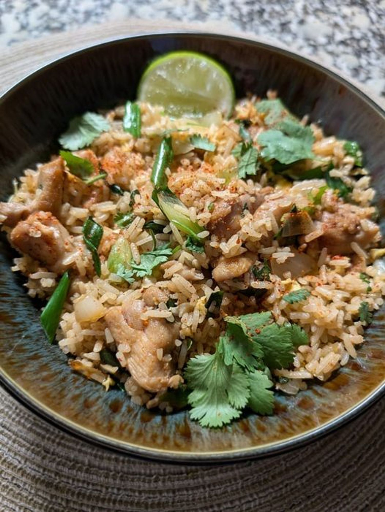 A black bowl with brown/blue interior with chicken fried rice w/onions, egg, green onions, lime and cilantro leaves.