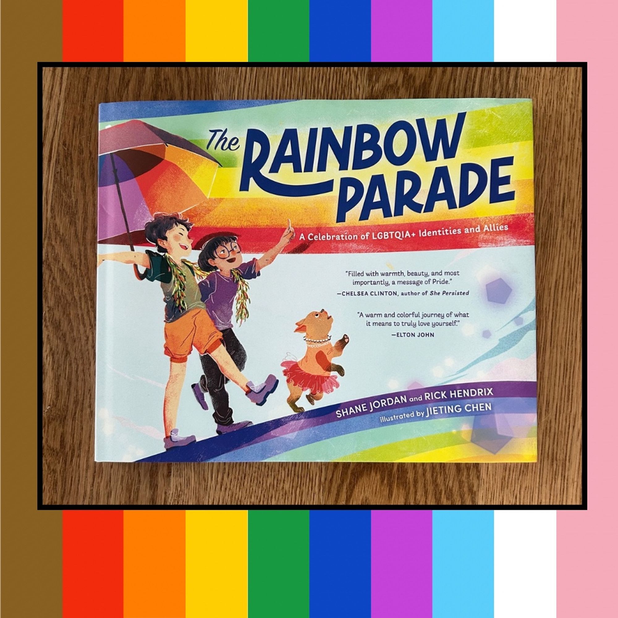 Graphic of a photo of the picture book THE RAINBOW PARADE: A CELEBRATION OF LGBTQIA+ IDENTITIES AND ALLIES by Shane Jordan and Rick Hendrix, illustrated by Jieting Chen. Image on the cover shows two children walking together under a rainbow umbrella, a boa draped around both their shoulders and a dog in a tutu.