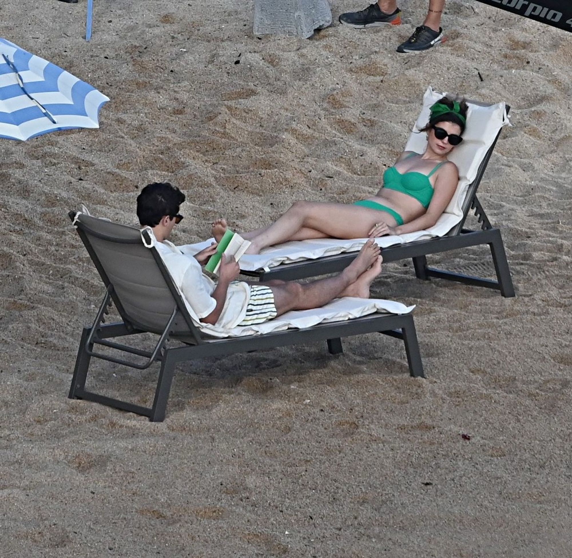 Imagem de Emily Bader e Tom Blyth sentados em cadeiras reclináveis na praia. Ele está lendo um livro.