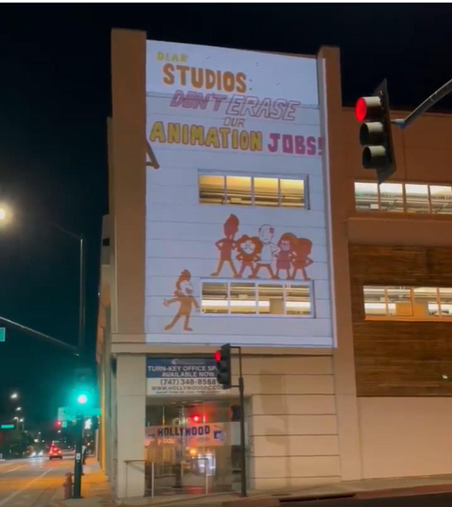 Photo of a building in downtown Burbank at night. An animated clip is projected on the side of the building. A group of people stand under text that reads "Dear studios: don't erase our animation jobs!"